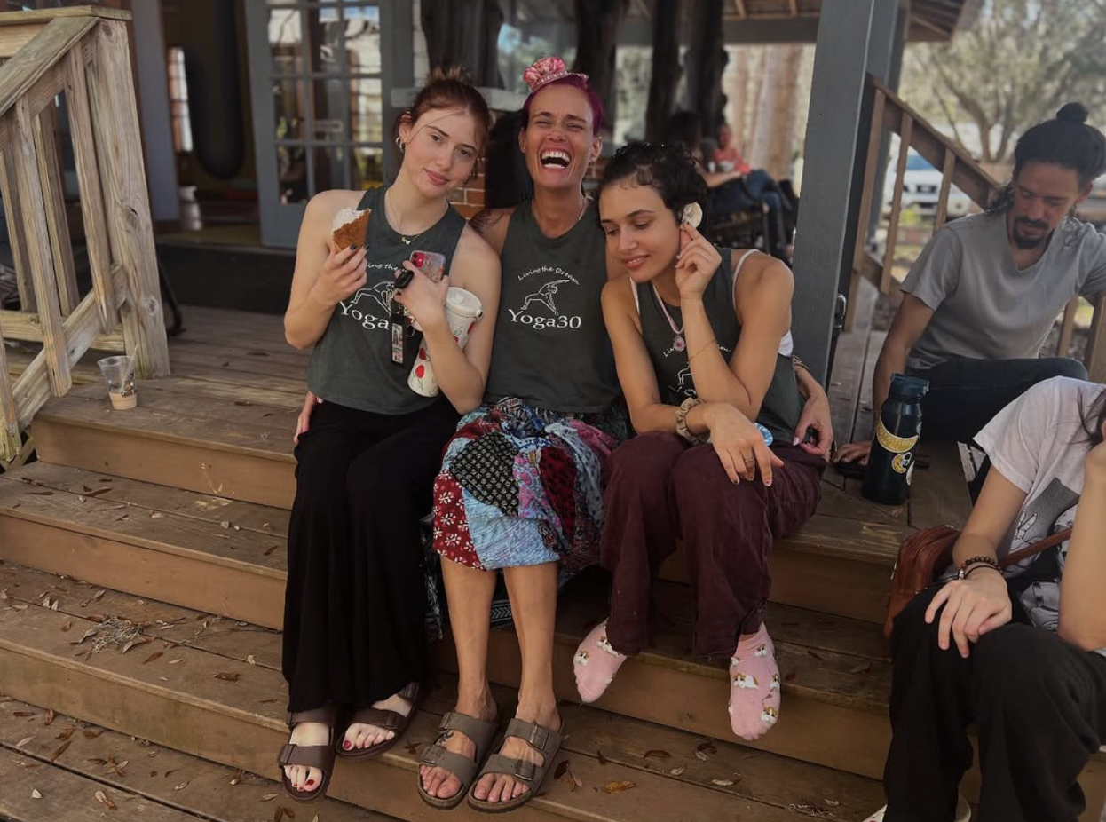 Three women wearing matching 'Yoga30' shirts sitting on wooden stairs outdoors, laughing and posing for a photo. Two women on the left are holding snacks, and a man is sitting on the right side of the stairs in the background.