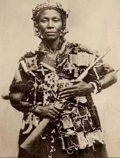 A woman in traditional attire with jewelry, holding a rifle and a stick.
