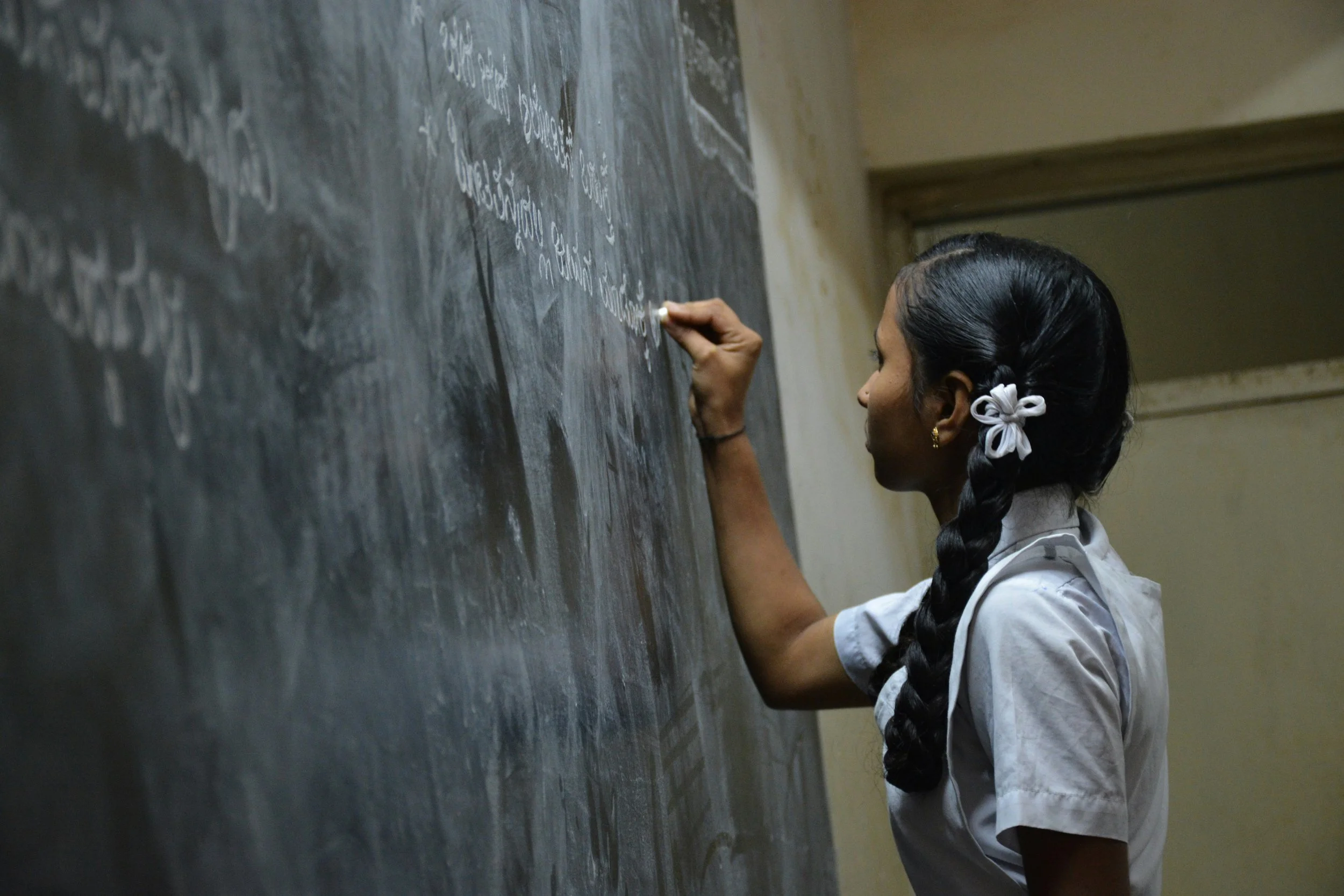 A girl with long braided hair and earrings writing on a blackboard with white chalk.