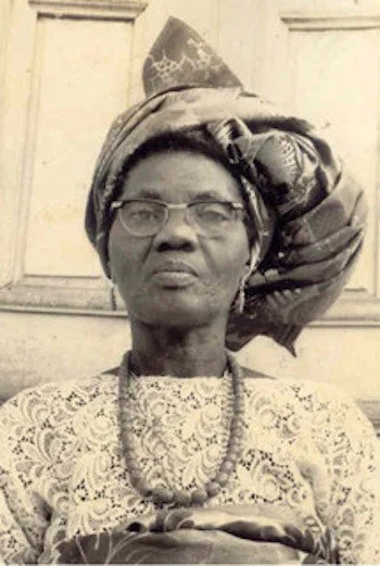 A woman wearing glasses, traditional headwrap, and jewelry, standing in front of a door.