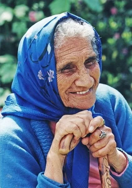 Close-up of elderly woman in blue headscarf smiling outdoors.