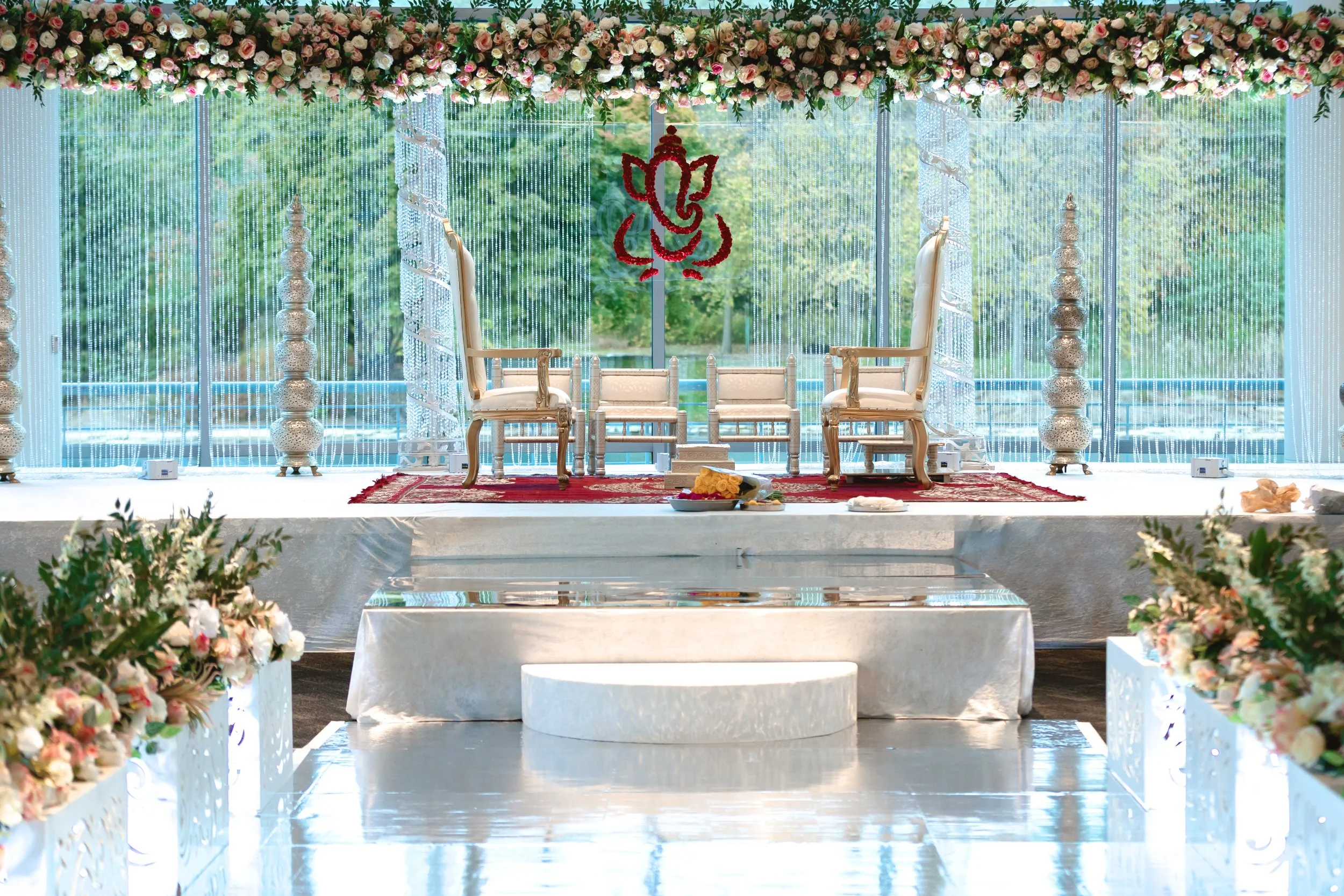 Wedding stage decorated with floral arrangements, two white ornate chairs for the couple, a red carpet, and a backdrop featuring the Hindu deity Ganesh and a divine elephant motif. The stage has a scenic view of greenery through large windows.