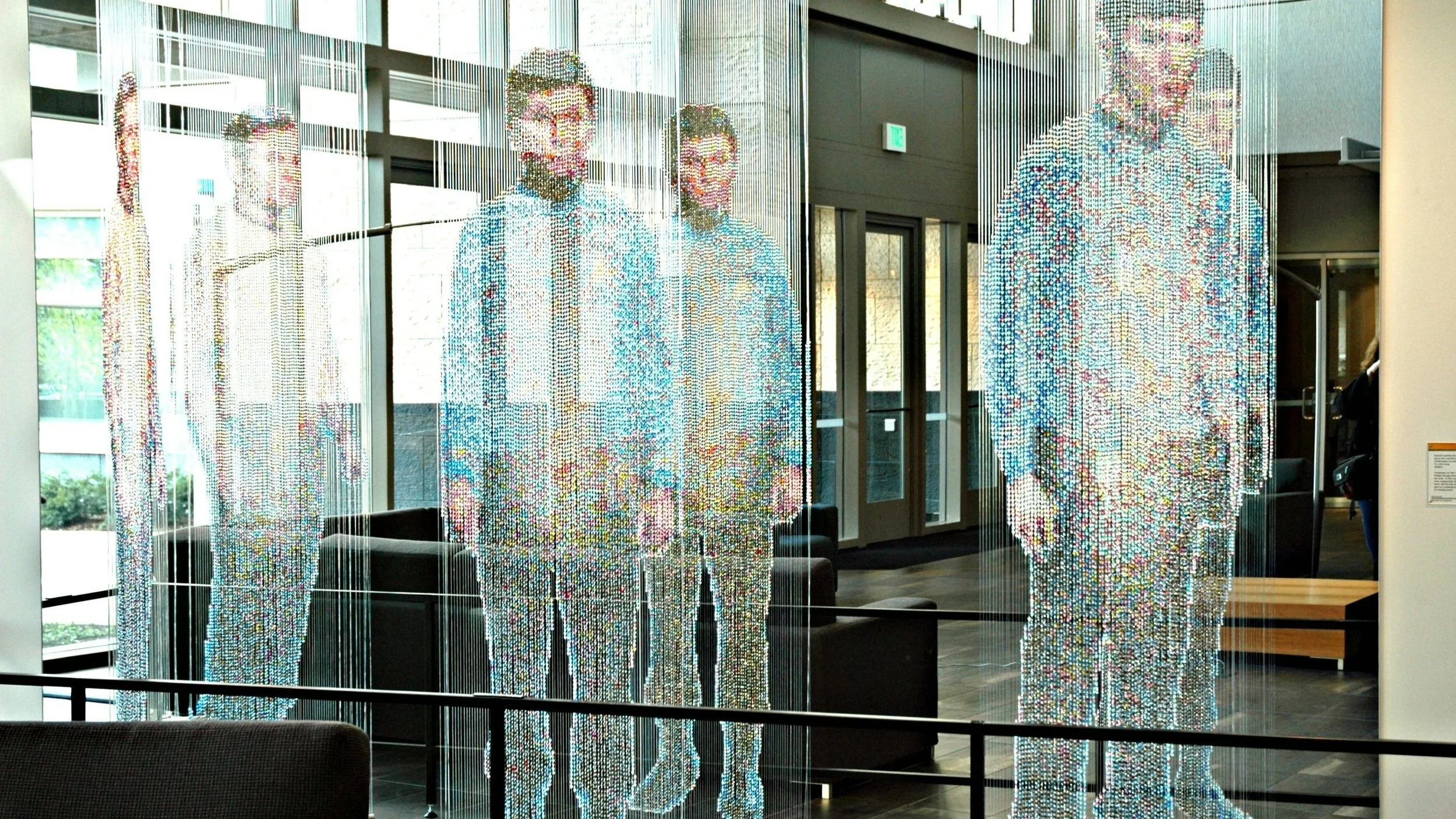 Kunstinstallation von Fotografie aus schwebenden Fäden, die bei Betrachtung aus einem bestimmten Winkel zu vollständigen menschlichen Figuren werden. Mehrere Personen erscheinen dreidimensional in einem modernen Innenraum, umgeben von großen Fenstern, mit gelbem Schild an der Wand.