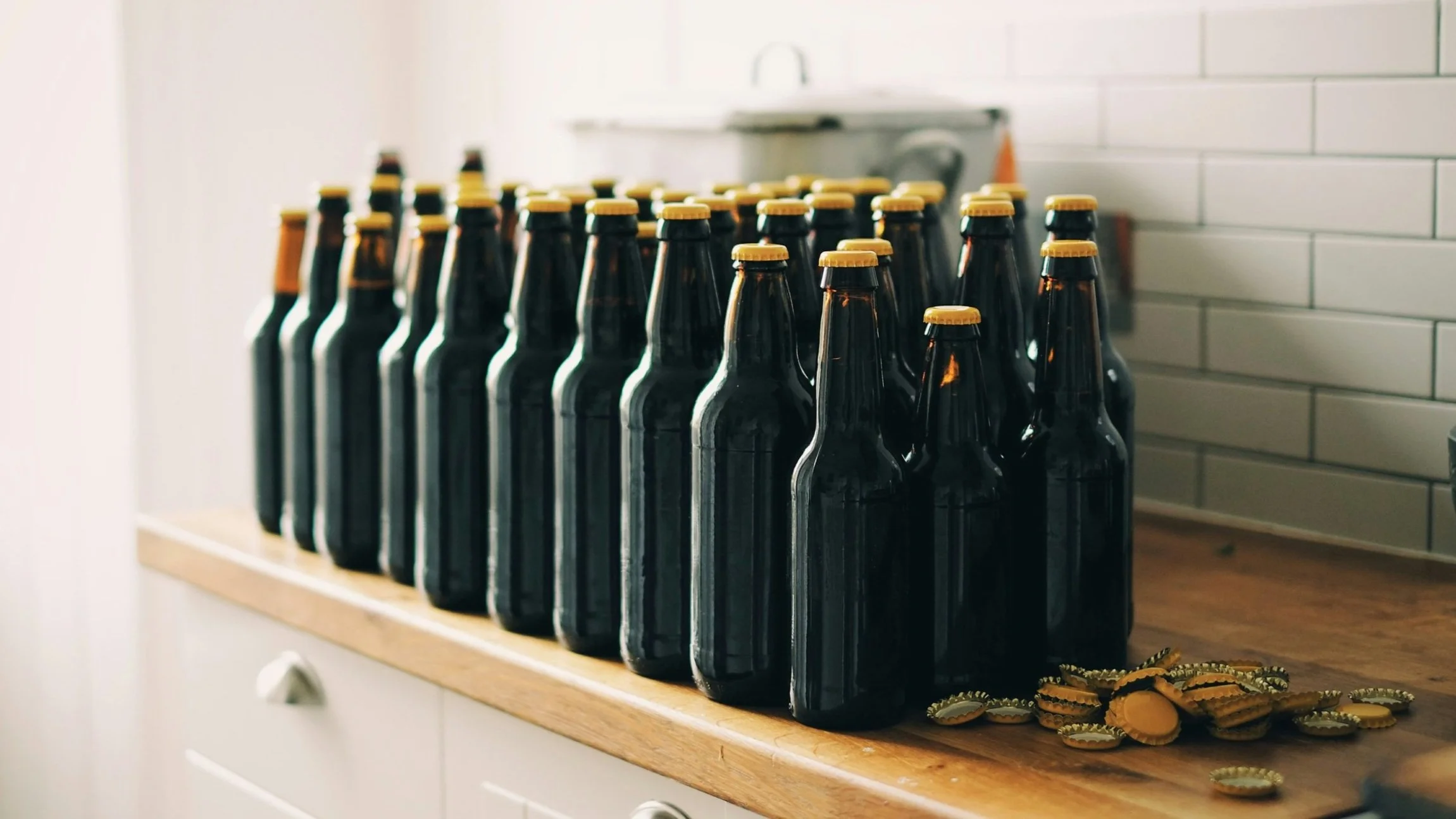 Reihe von dunklen Bierflaschen mit goldenen Kronkorken auf einem Holztisch, daneben lose Kronkorken.