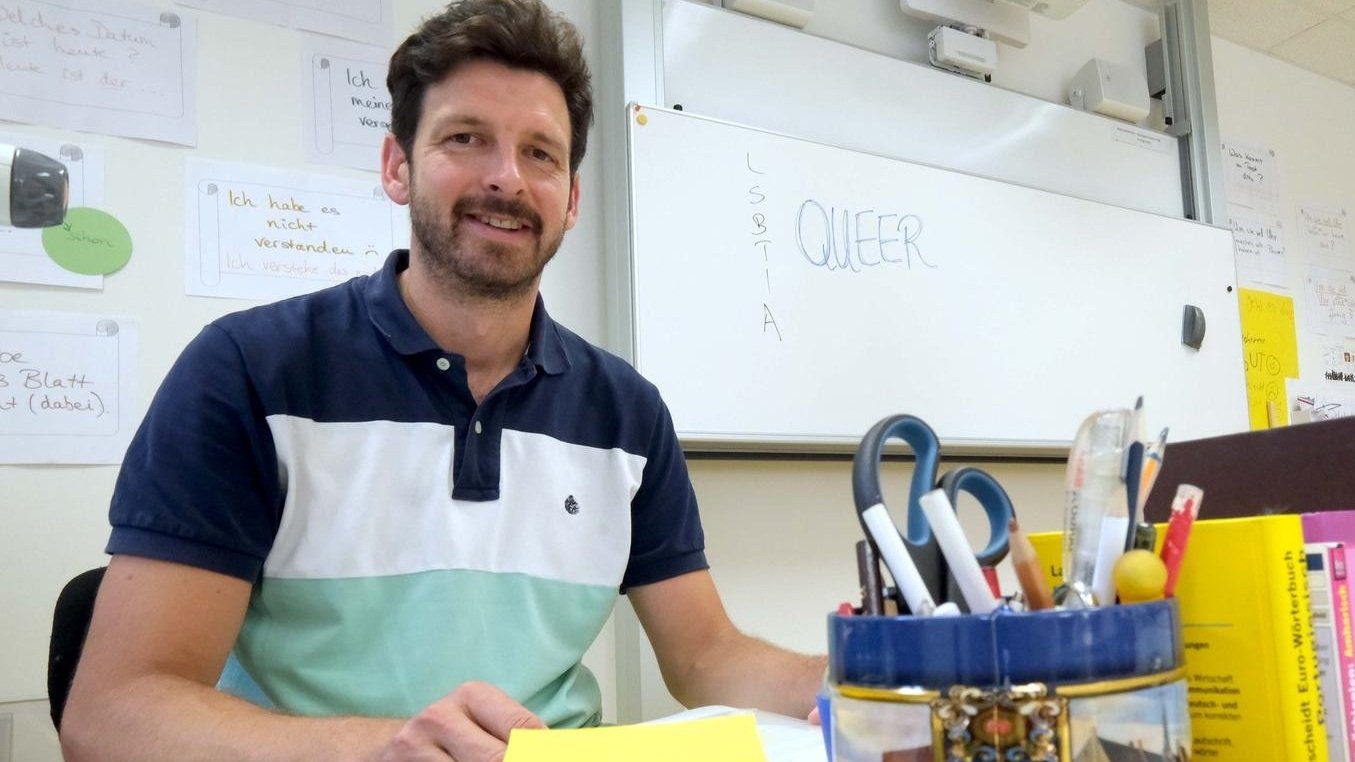 Ein Mann sitzt an einem Tisch in einem Klassenzimmer, lächelt in die Kamera. Im Hintergrund ist ein Whiteboard mit den Worten „QUEER“ und „LSBTIA“ zu sehen. Auf dem Tisch stehen Schreibutensilien und Bücher.