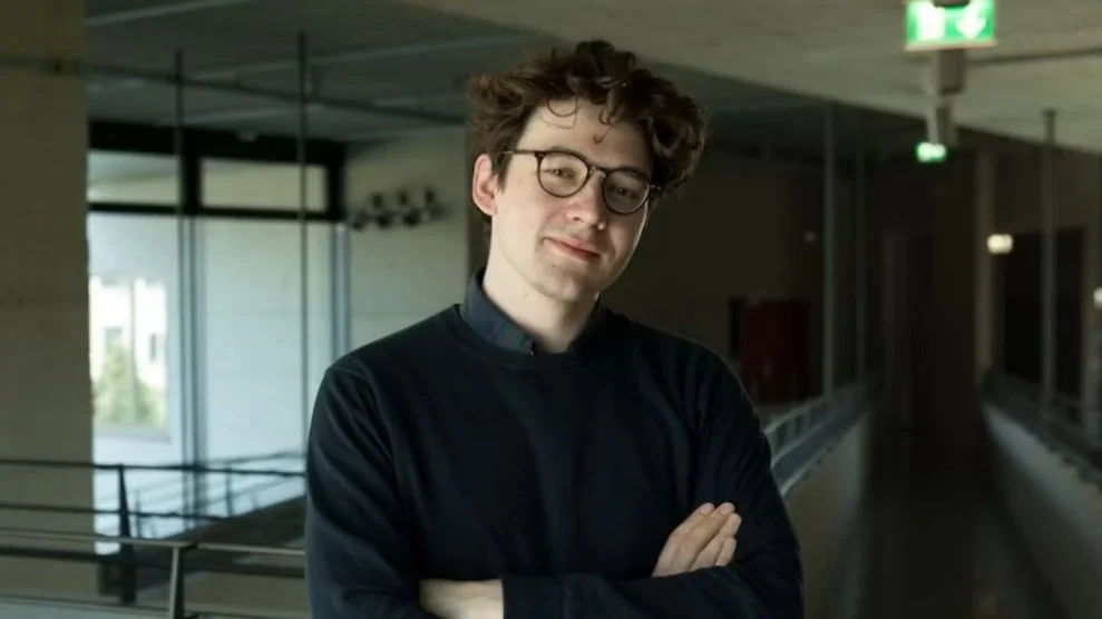 Mann mit Brille im Flur eines modernen Gebäudes, mit verschränkten Armen, lächelt leicht.