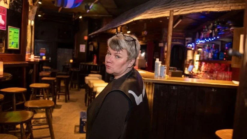 Innenansicht einer Bar mit Holztischen und -stühlen, ein Mann mit Brille auf dem Kopf dreht sich zur Kamera um, Bar im Hintergrund mit Getränken.