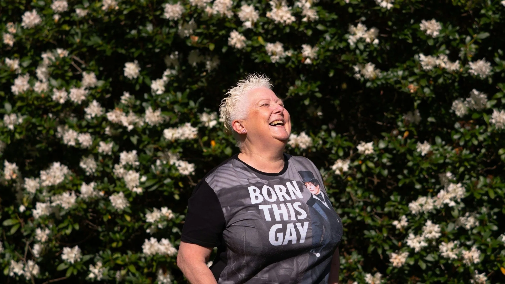 Lachende Person mit kurzem, blondem, stacheligem Haar trägt ein T-Shirt mit der Aufschrift „Born This Gay“ vor einem Hintergrund aus grünen Blättern und weißen Blumen.