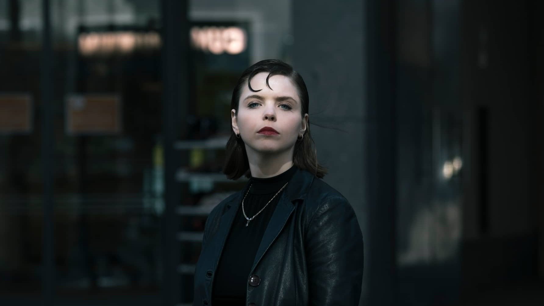 Frau mit kurzem, dunklem Haar in schwarzer Lederjacke, posiert ernsthaft vor unscharfem Hintergrund.