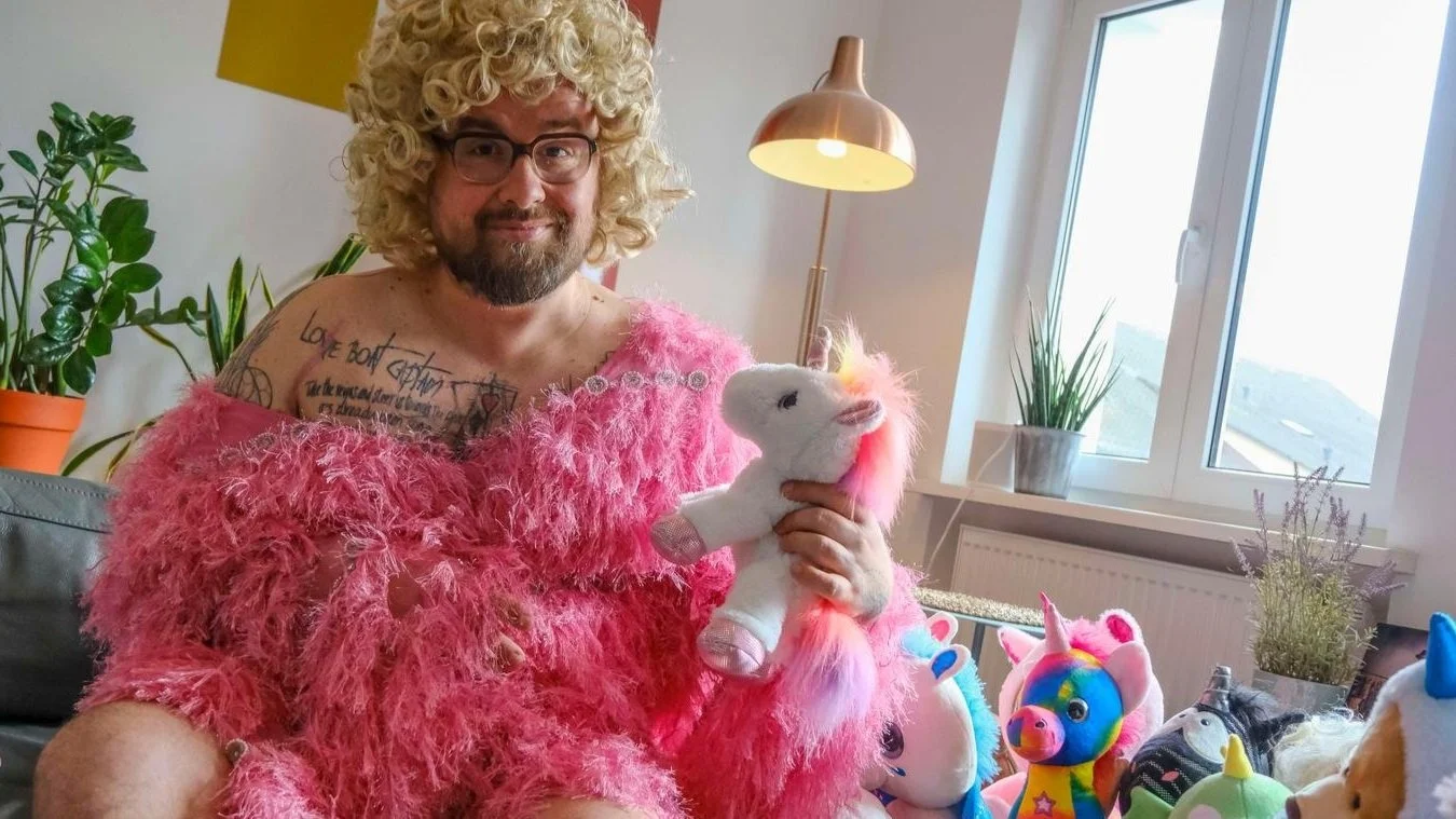 Mann mit blonder Perücke und rosa Federkleid, hält ein Plüscheinhorn. Hintergrund zeigt Pflanzen und ein Fenster.