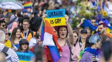 Menschen bei einer LGBTQ+ Parade, halten Regenbogenflaggen und Schilder. Ein Schild trägt die Aufschrift "Ukraine is Gay". Die Stimmung ist fröhlich und bunt.