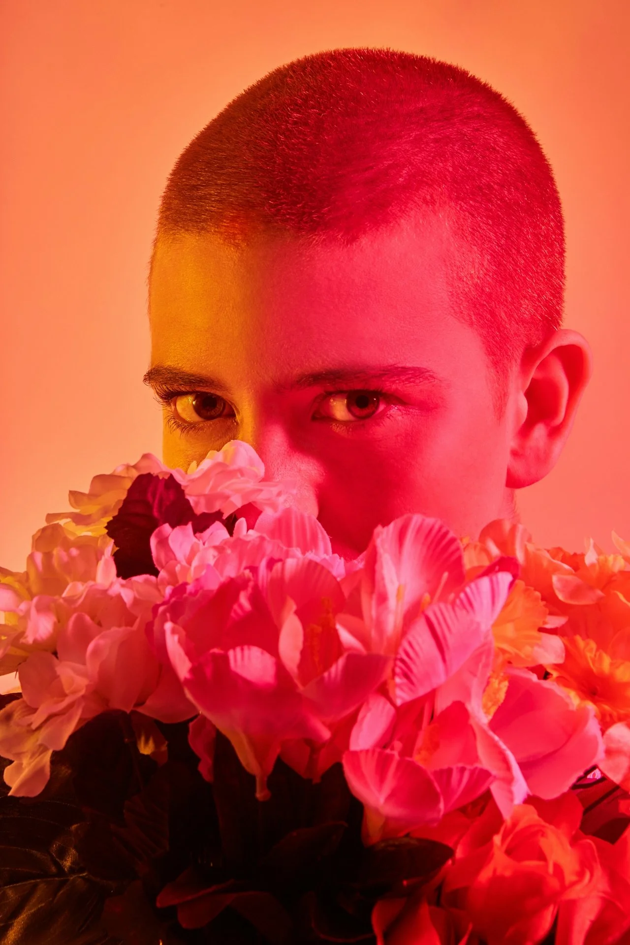 Person with buzz cut partially obscured by colorful flowers under red-orange lighting.
