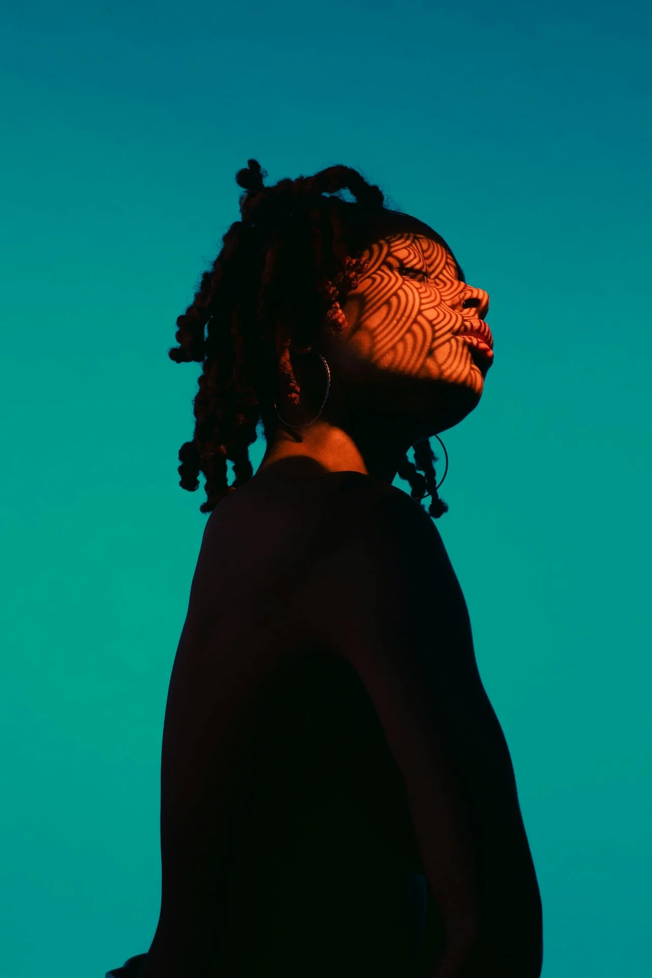A woman with dreadlocks wearing hoop earrings, illuminated by intricate shadow patterns on her face, against a teal background.