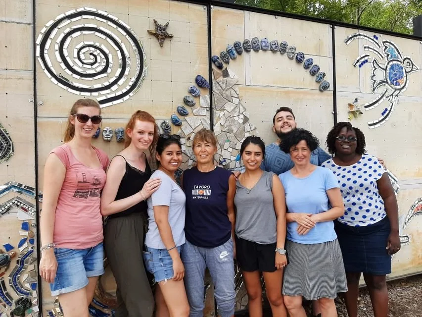 Group of eight people smiling in front of a mosaic wall with various designs, including a spiral and abstract shapes.