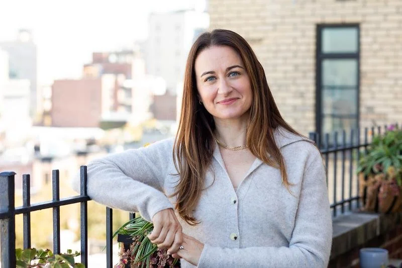 A woman with long brown hair, wearing a light gray sweater and a gold necklace, leaning on a balcony railing with a cityscape in the background.