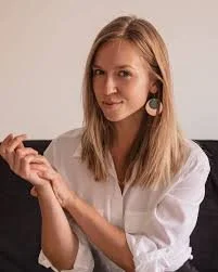 Young woman with blonde hair wearing a white shirt and large earrings, holding a phone and looking at the camera.