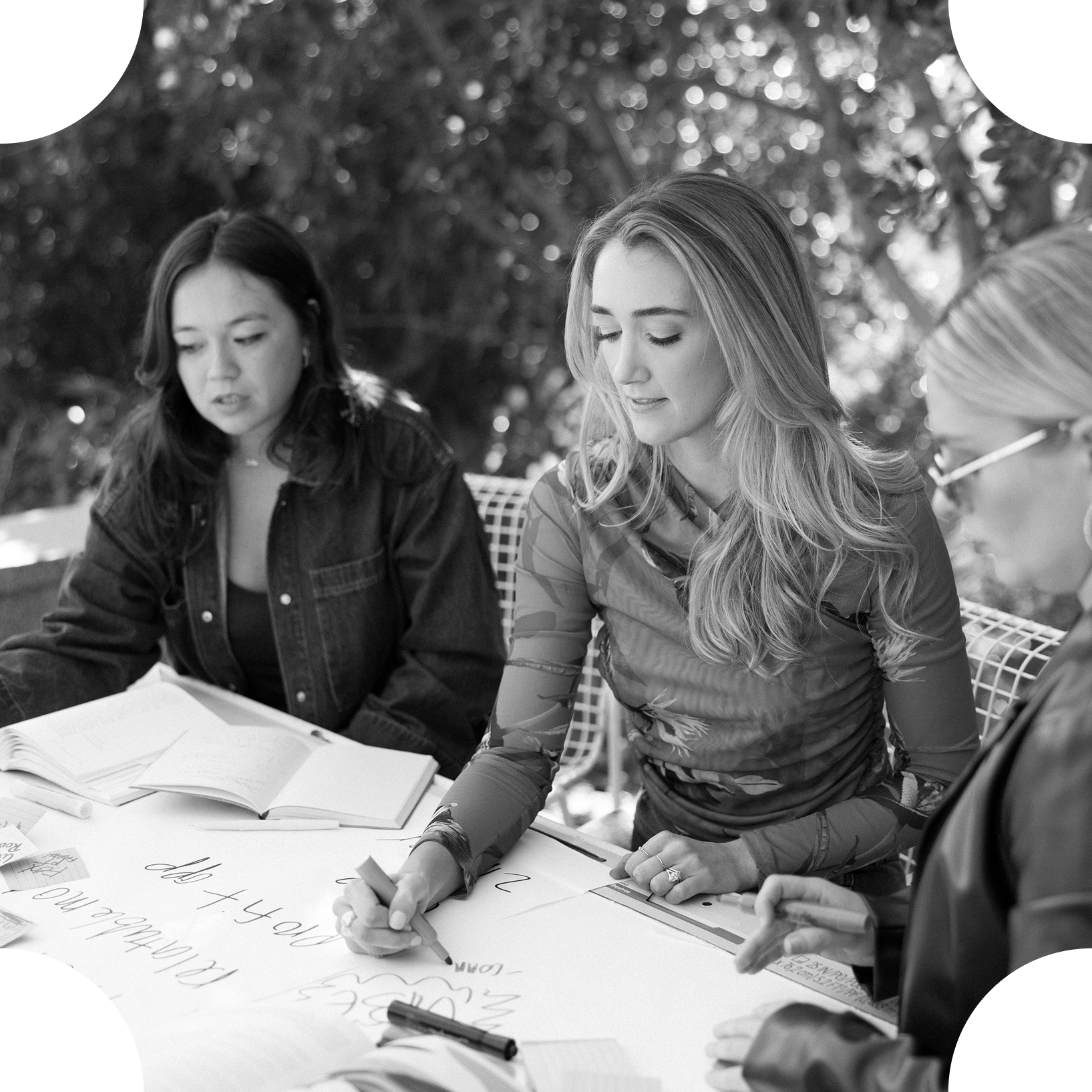 Three women sit around a table outdoors, engaged in a discussion or collaborative activity, with notebooks and papers on the table.