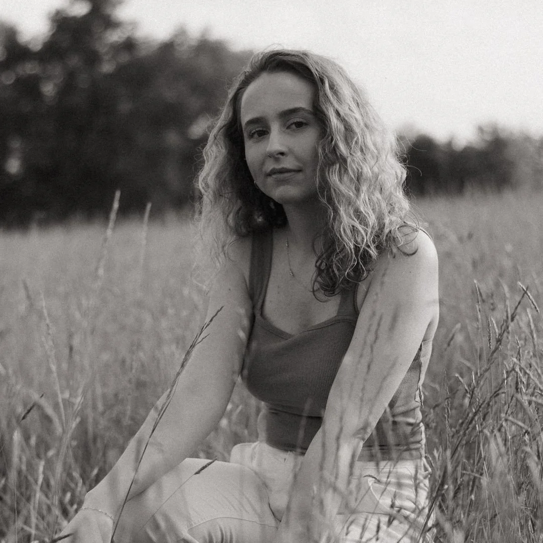 Black and white photo of a woman sitting in a field of tall grass, looking at the camera with a calm expression.