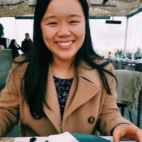 Young woman smiling, sitting at a restaurant table in a casual setting with large windows and other patrons in the background.