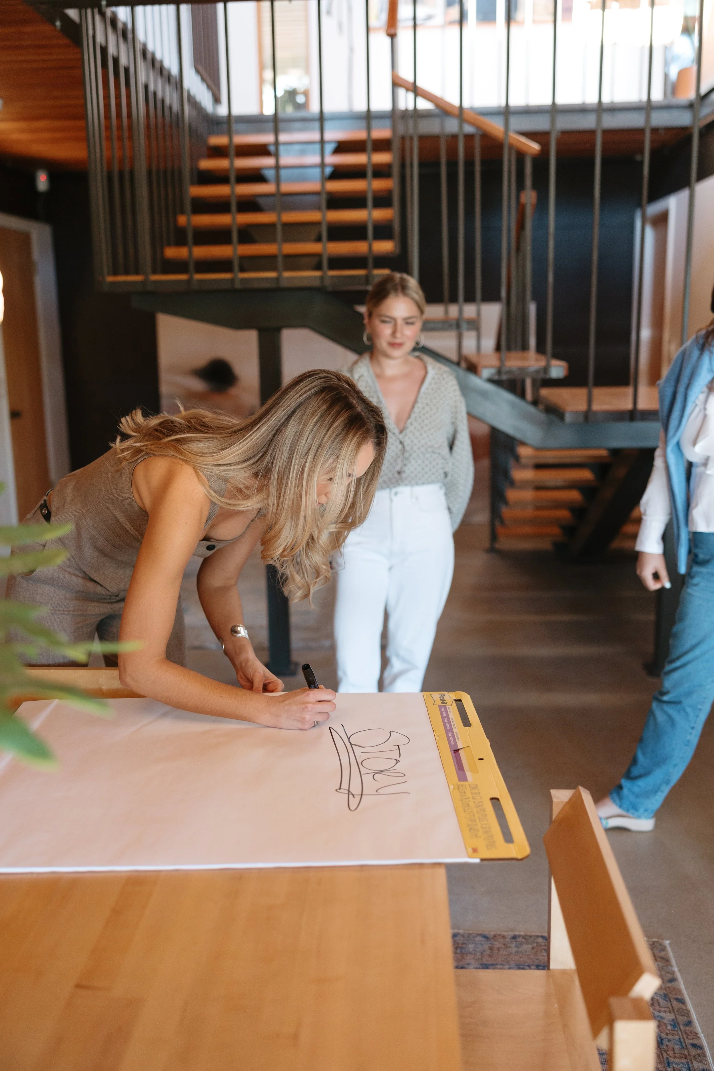 A woman is signing a large card or poster on a table, with two other women watching her in a modern interior with a staircase in the background.