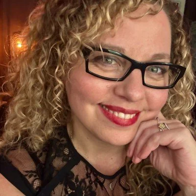 Close-up photo of a woman with curly blond hair, black glasses, and red lipstick, smiling and resting her chin on her hand.