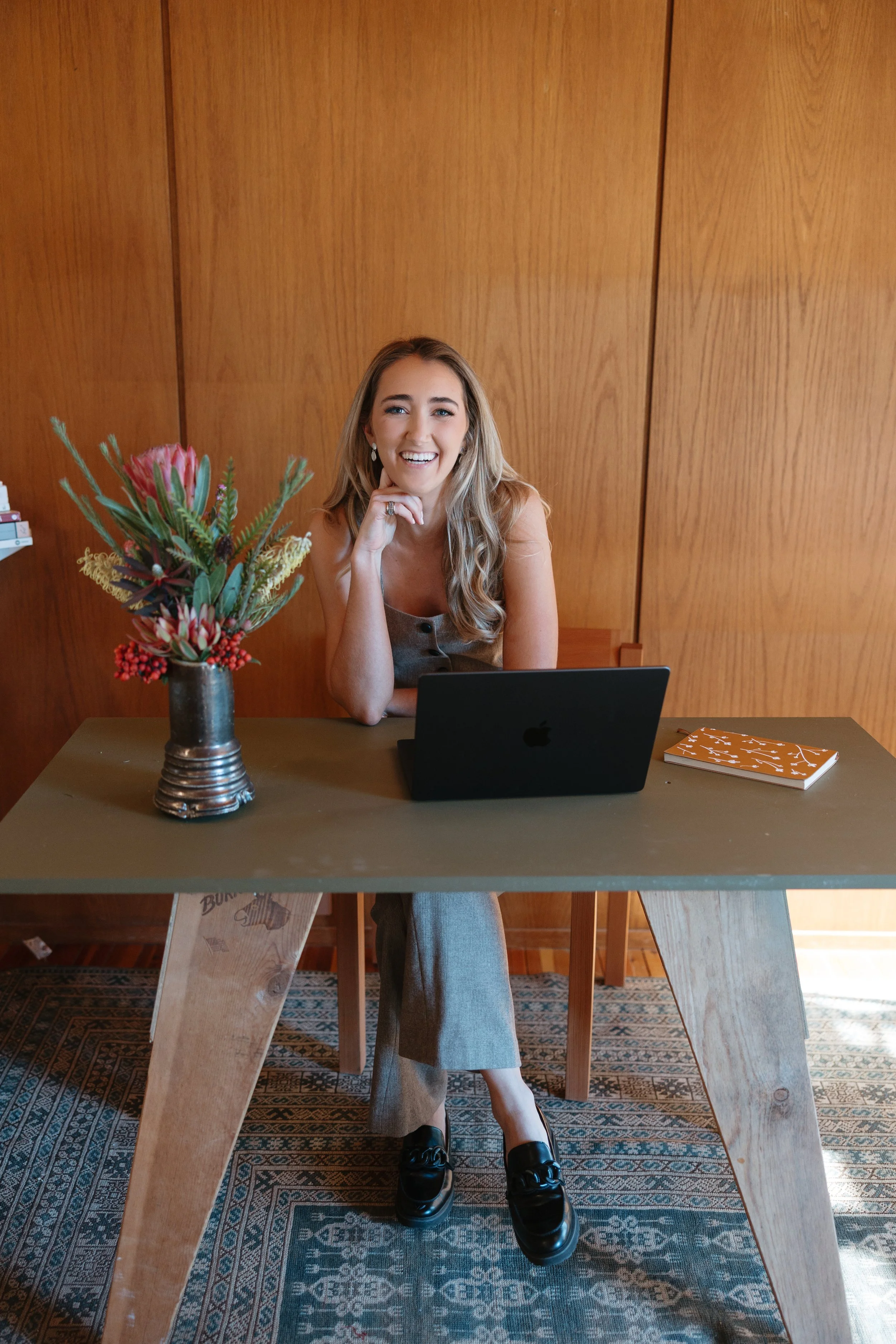 A woman with long blonde hair smiling, sitting at a desk with a laptop, decorated with a vase of flowers, a notebook, and a wooden wall background.