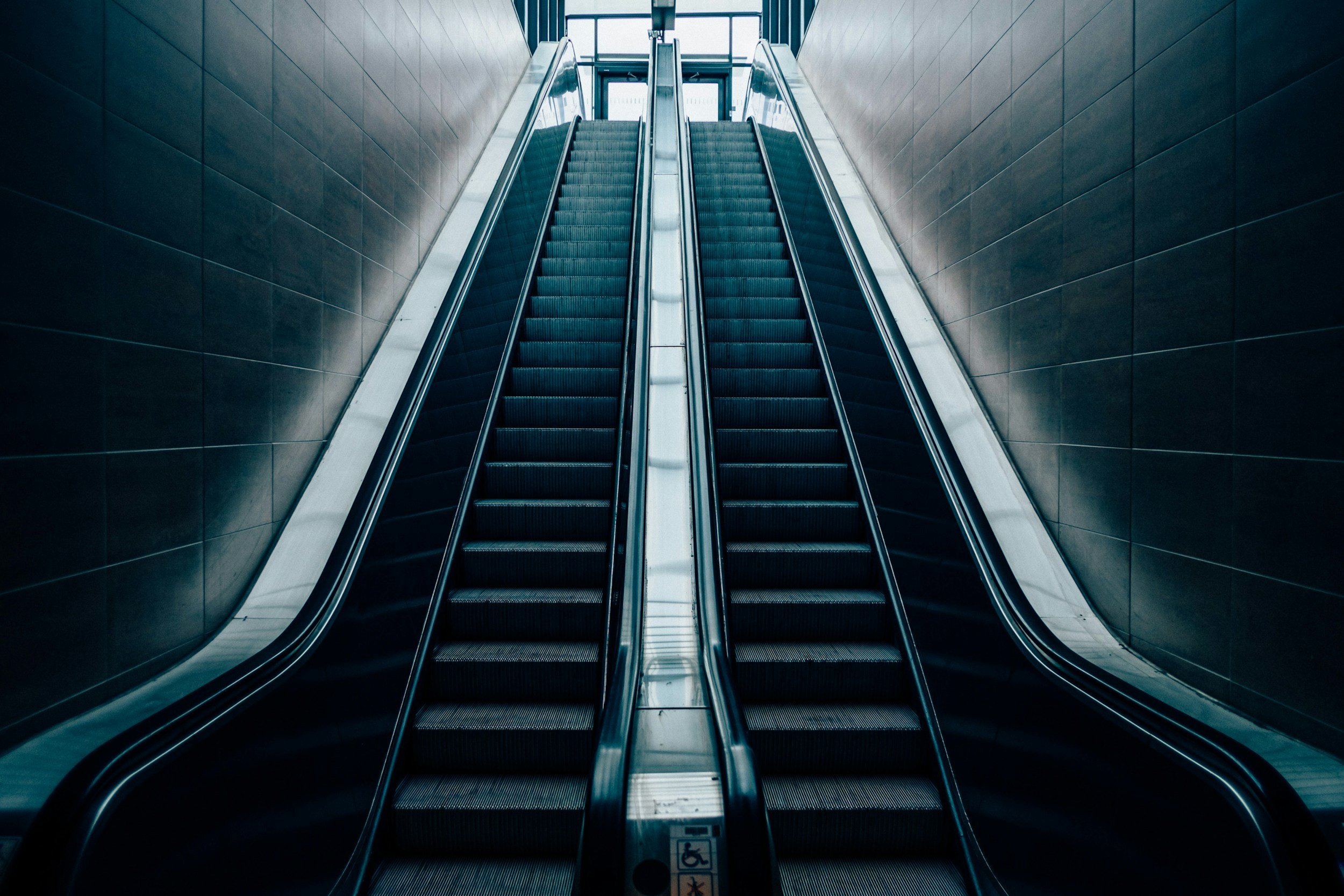 Indoor escalators ascending between dark tiled walls with glass windows at the top.