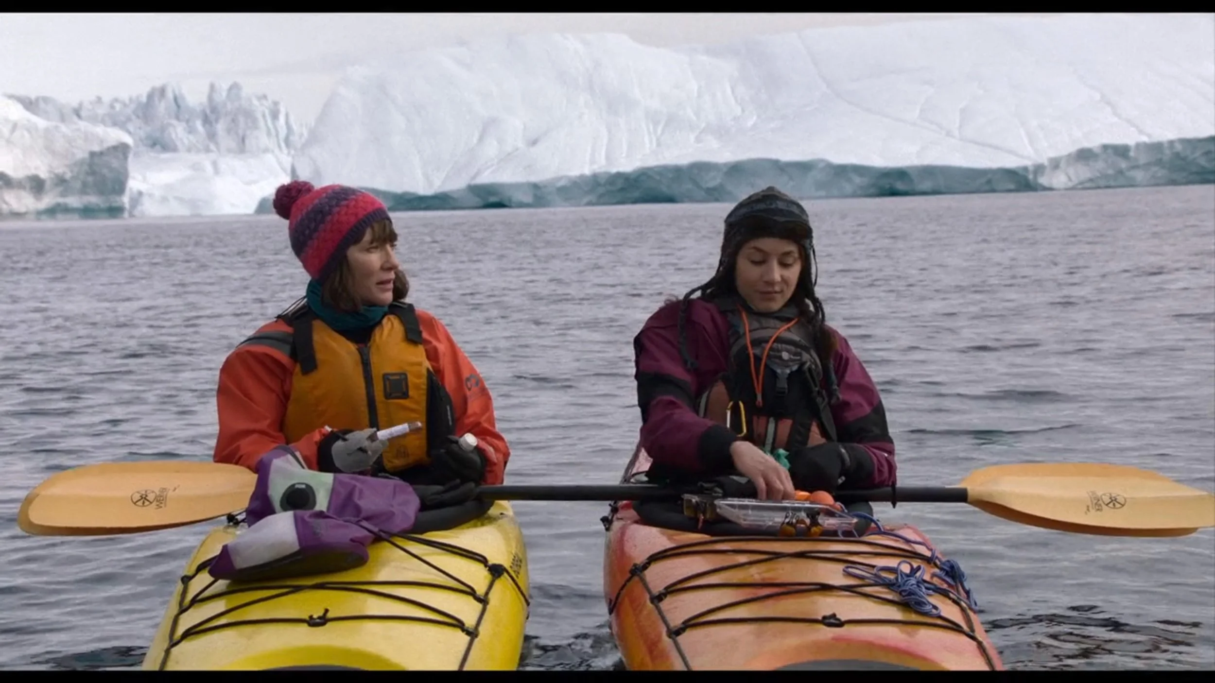 Two women kayaking on a lake with icebergs in the background, wearing warm gear and holding paddles, one in an orange life jacket, the other in a purple jacket.