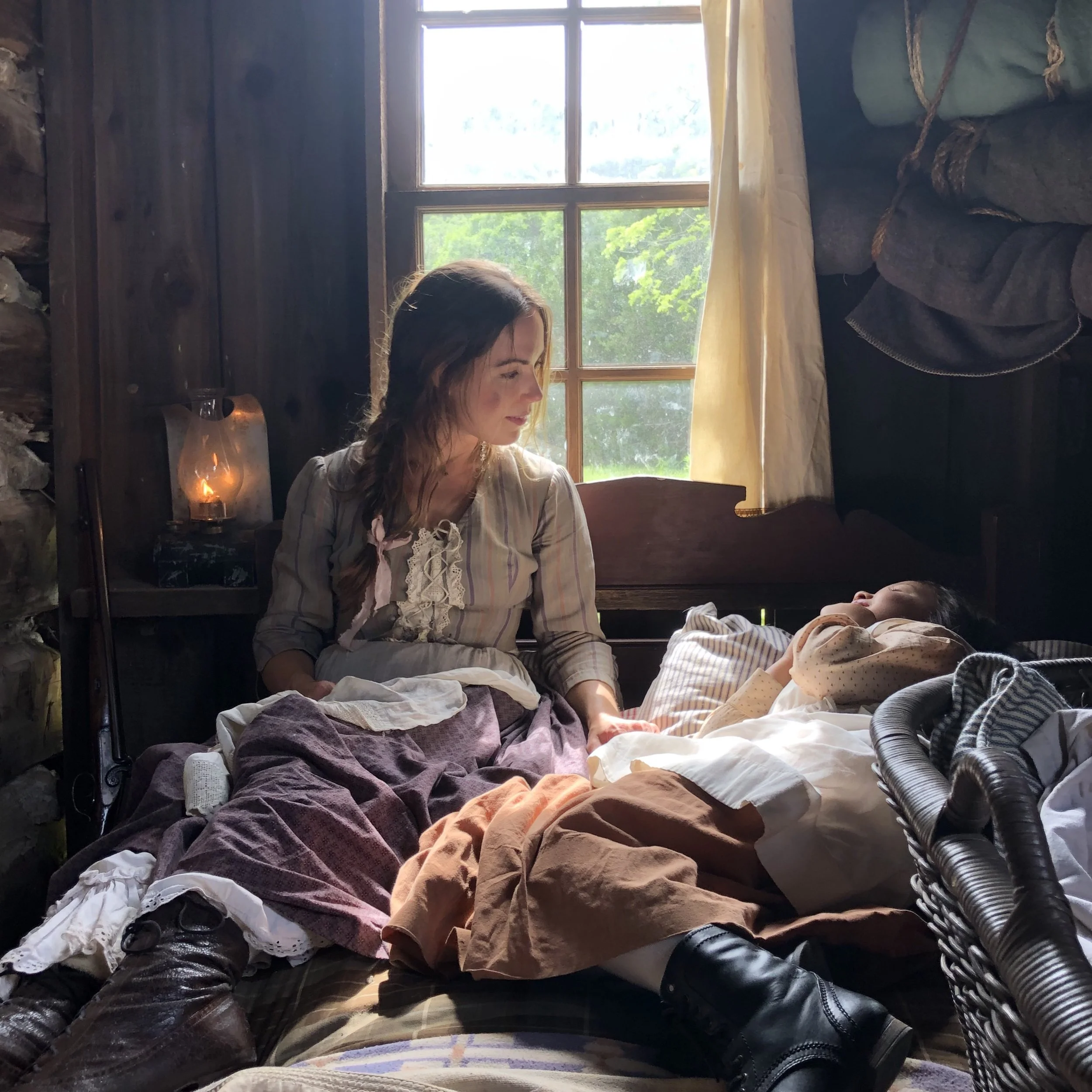A woman in historical clothing sits on a bed next to a child in a rustic wooden cabin, with light streaming through a window.