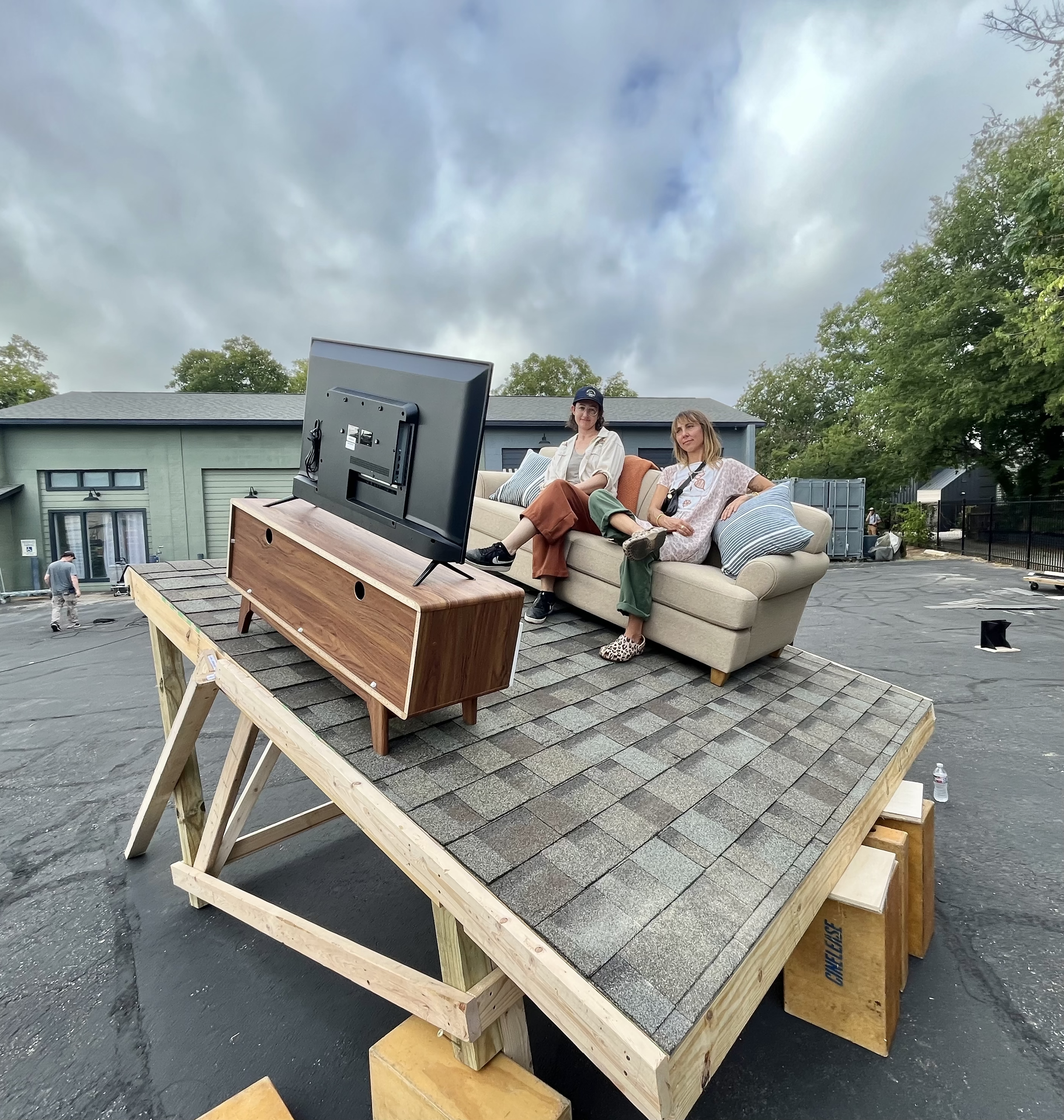 Two women sitting on a couch positioned on a tilted roof platform, facing a television on a wooden stand outdoors.
