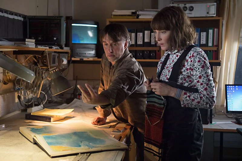 Two people working in a cluttered office with maps and files, discussing something on a large map spread out on a table, surrounded by dim lighting and computer monitors in the background.