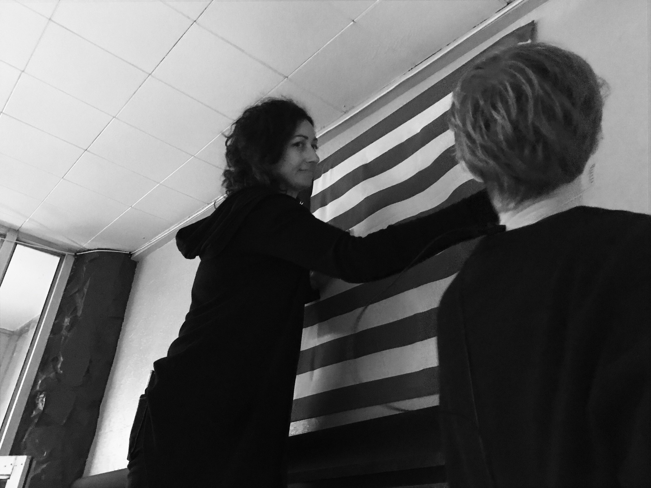 Two people hanging a striped fabric on a wall in a room with white ceiling tiles. The image is in black and white.
