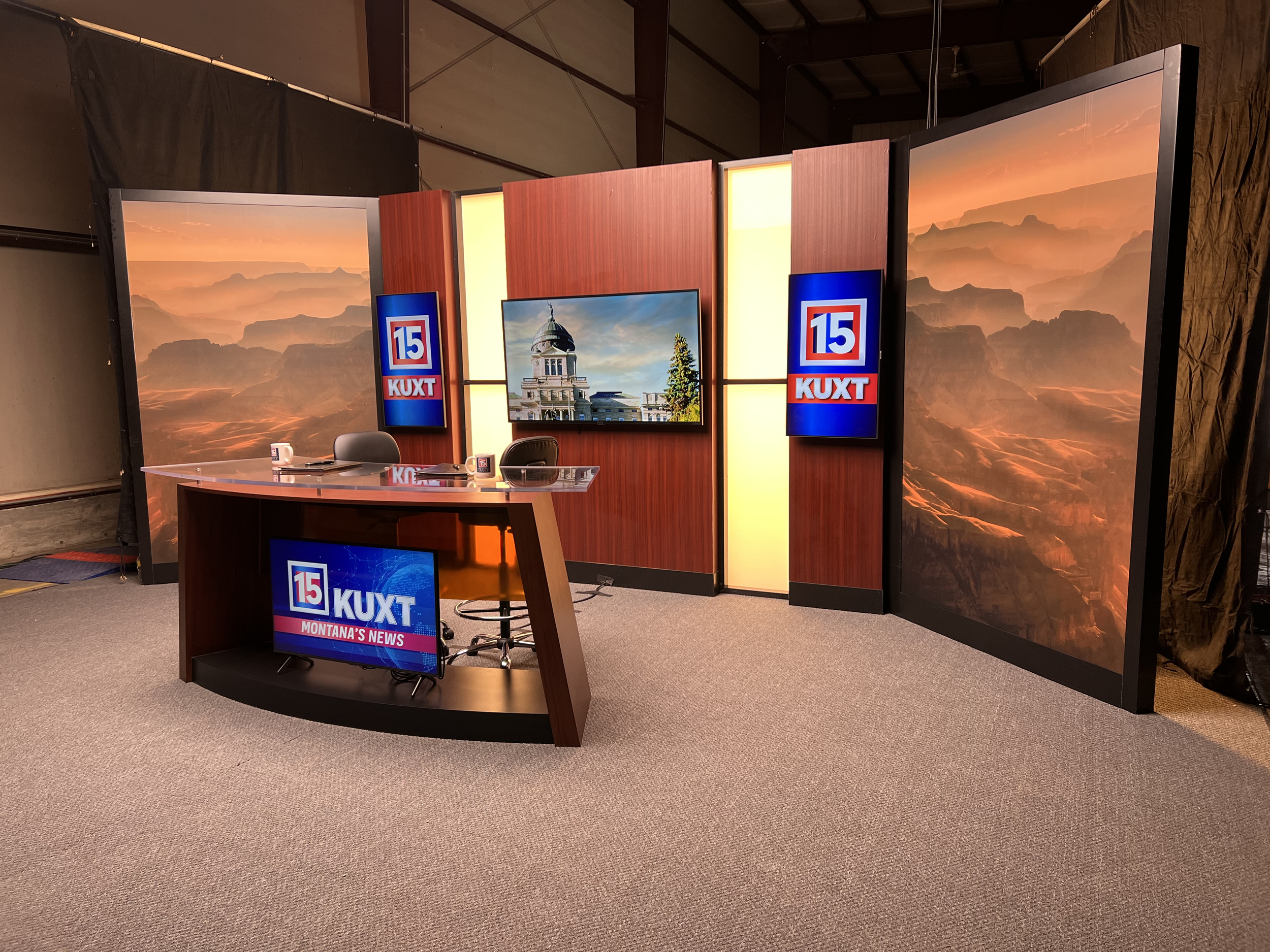 A television news studio setup with a wooden desk and chairs. The backdrop features panoramic images and has a large screen displaying "15 KUXT Montana's News." The set is arranged for broadcasting, with studio lights visible above.