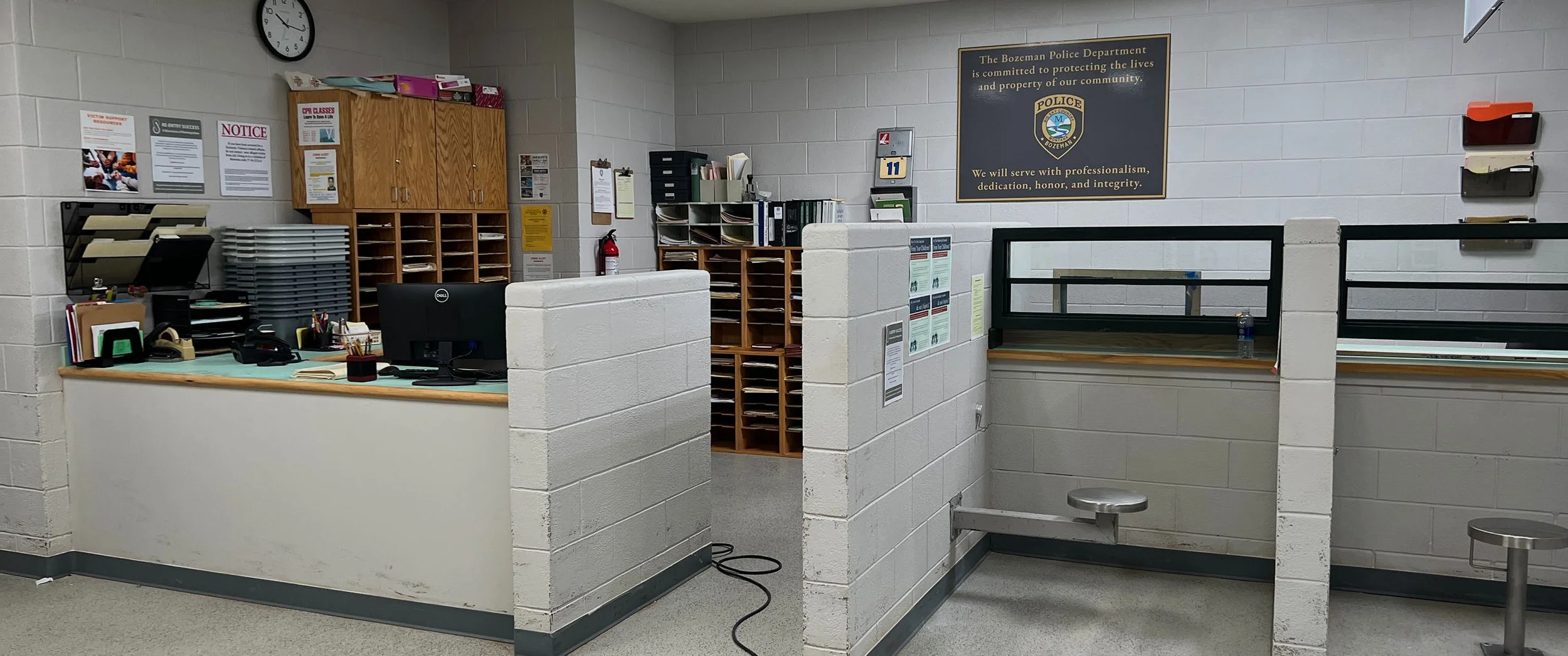 Office reception area in a police department with desks, office supplies, filing cabinets, a wall clock, and a sign displaying values and commitment of the department.