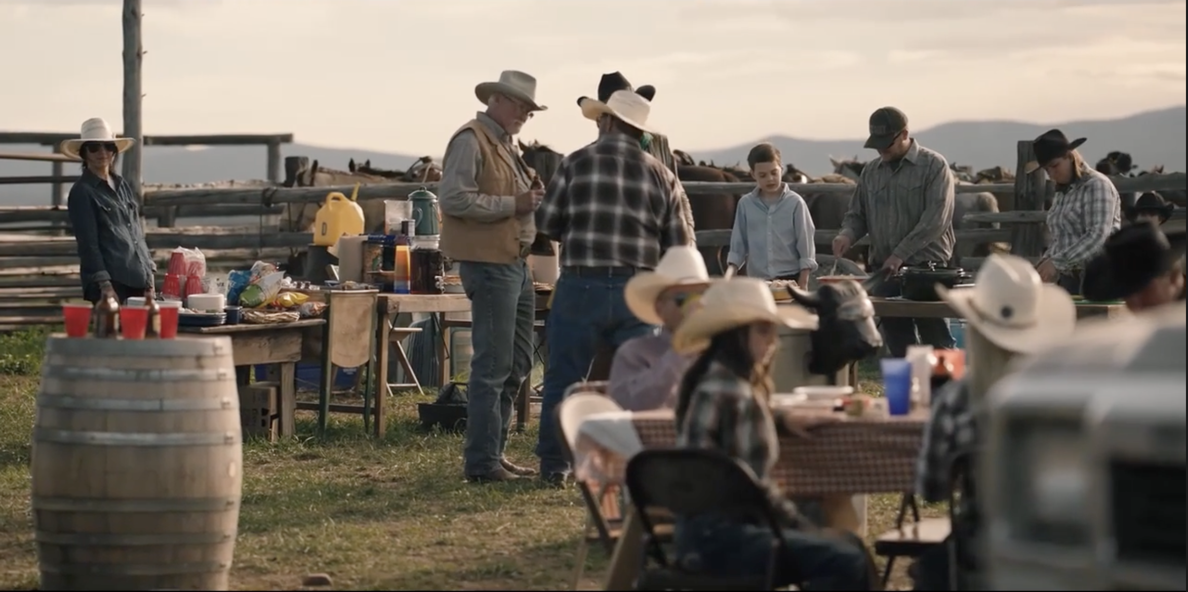 Outdoor gathering with people in cowboy hats and western attire, near a table with food and drinks. A wooden barrel with beer bottles is in the foreground.