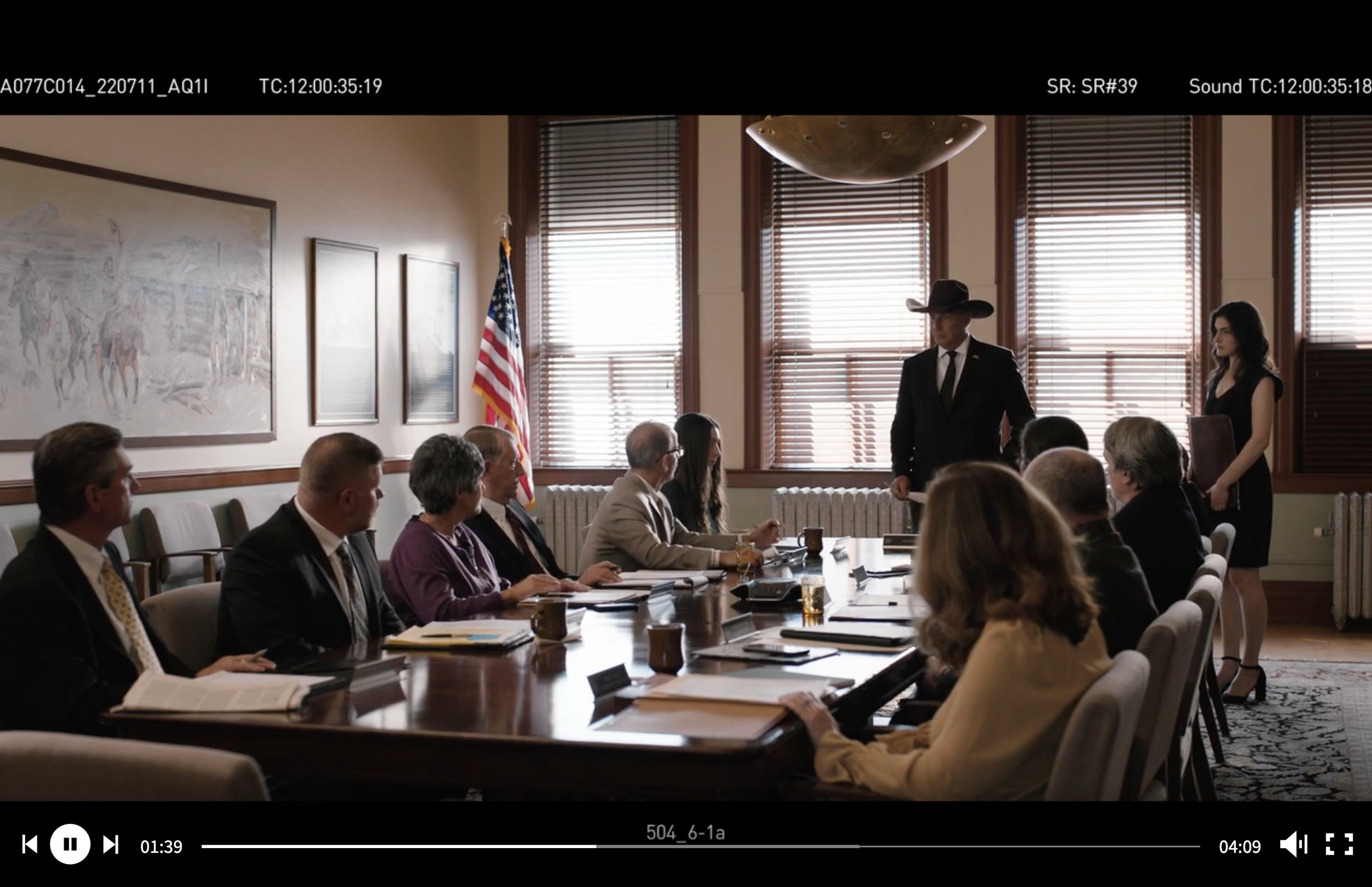 A group of people sitting at a conference table in a meeting room. One man, wearing a cowboy hat, is standing, and a woman is standing beside him holding a folder. An American flag is visible near the window with blinds, and paintings are on the wall