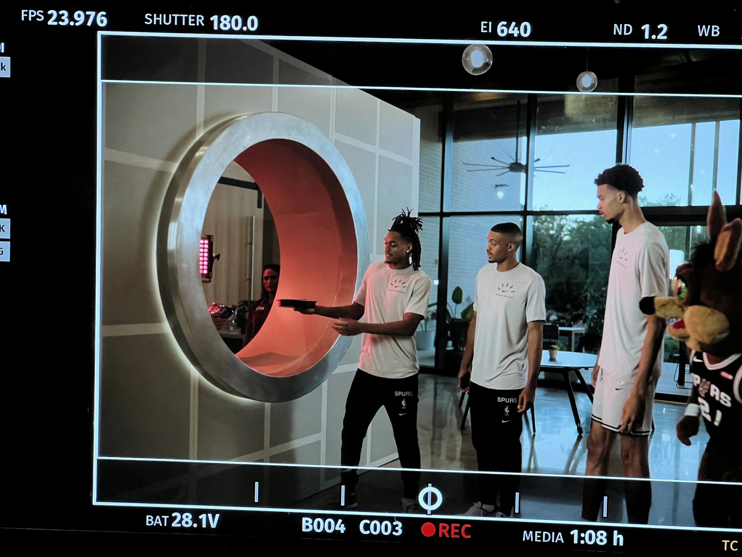 A behind-the-scenes shot of a video production showing three people in matching Spurs outfits interacting with a large circular structure. A person in a sports mascot costume is visible beside them. The production screen interface is visible with var