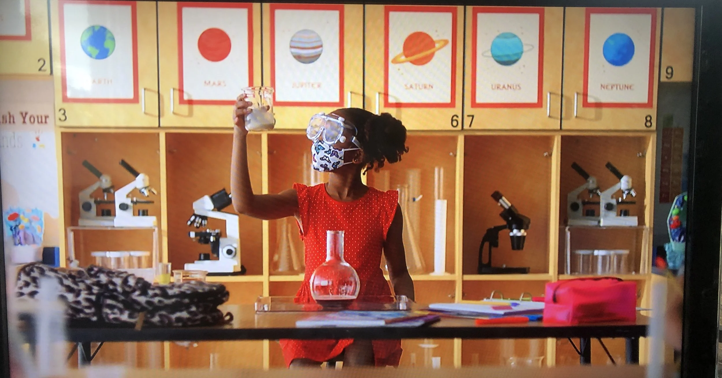 A child in a red dress and protective goggles holds a beaker in a classroom with educational posters of planets and microscopes on shelves.