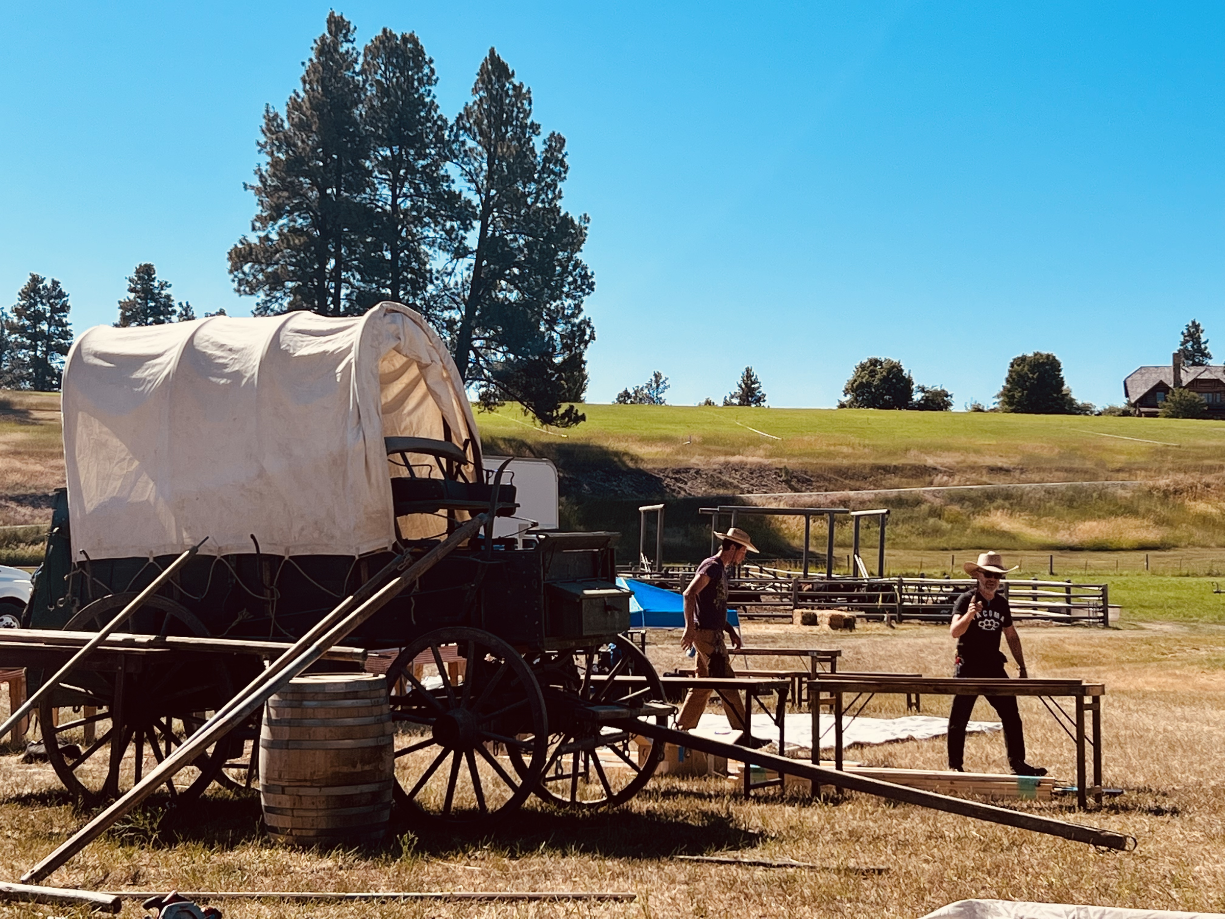 Two people setting up beside a covered wagon in a rural area with wooden structures and scattered trees in the background.