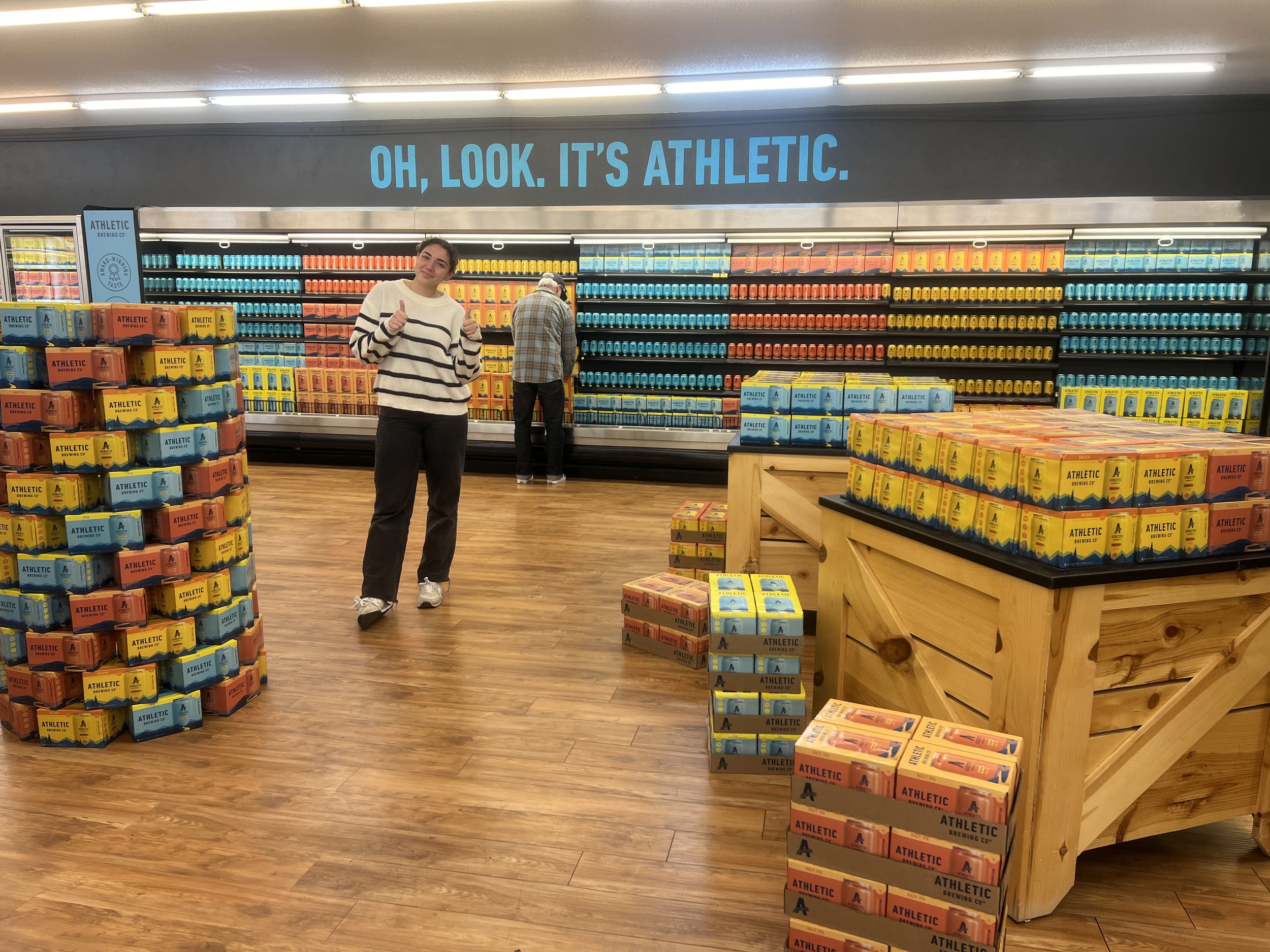 Person in a store posing in front of shelves and stacks of canned drinks from Athletic Brewing Company, with a sign that reads "Oh, Look. It's Athletic."
