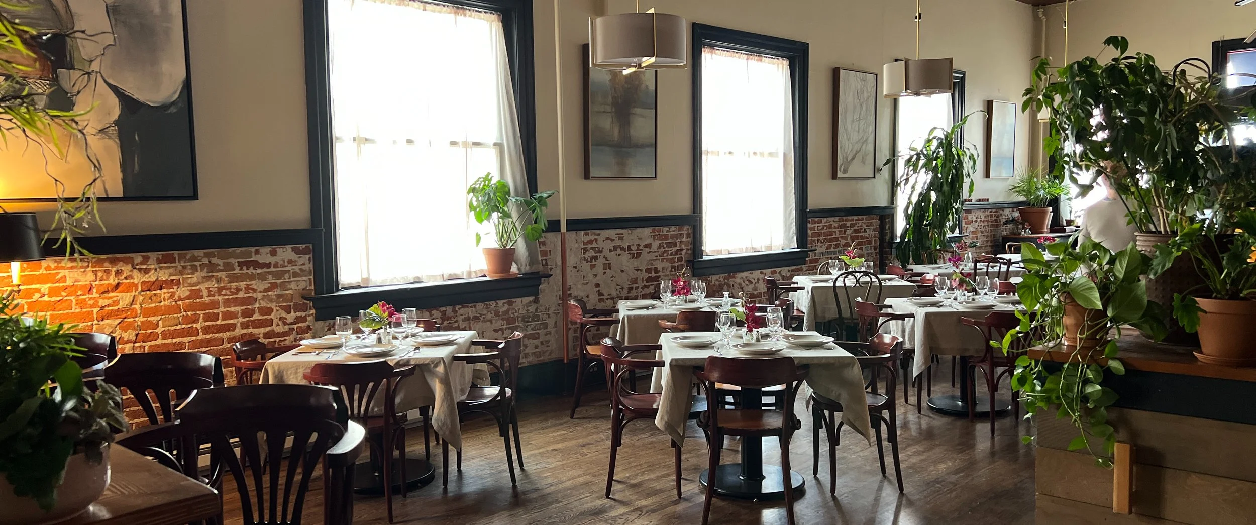 Cozy restaurant interior with brick walls, wooden flooring, and potted plants, featuring several tables with white tablecloths and set with plates and glasses.