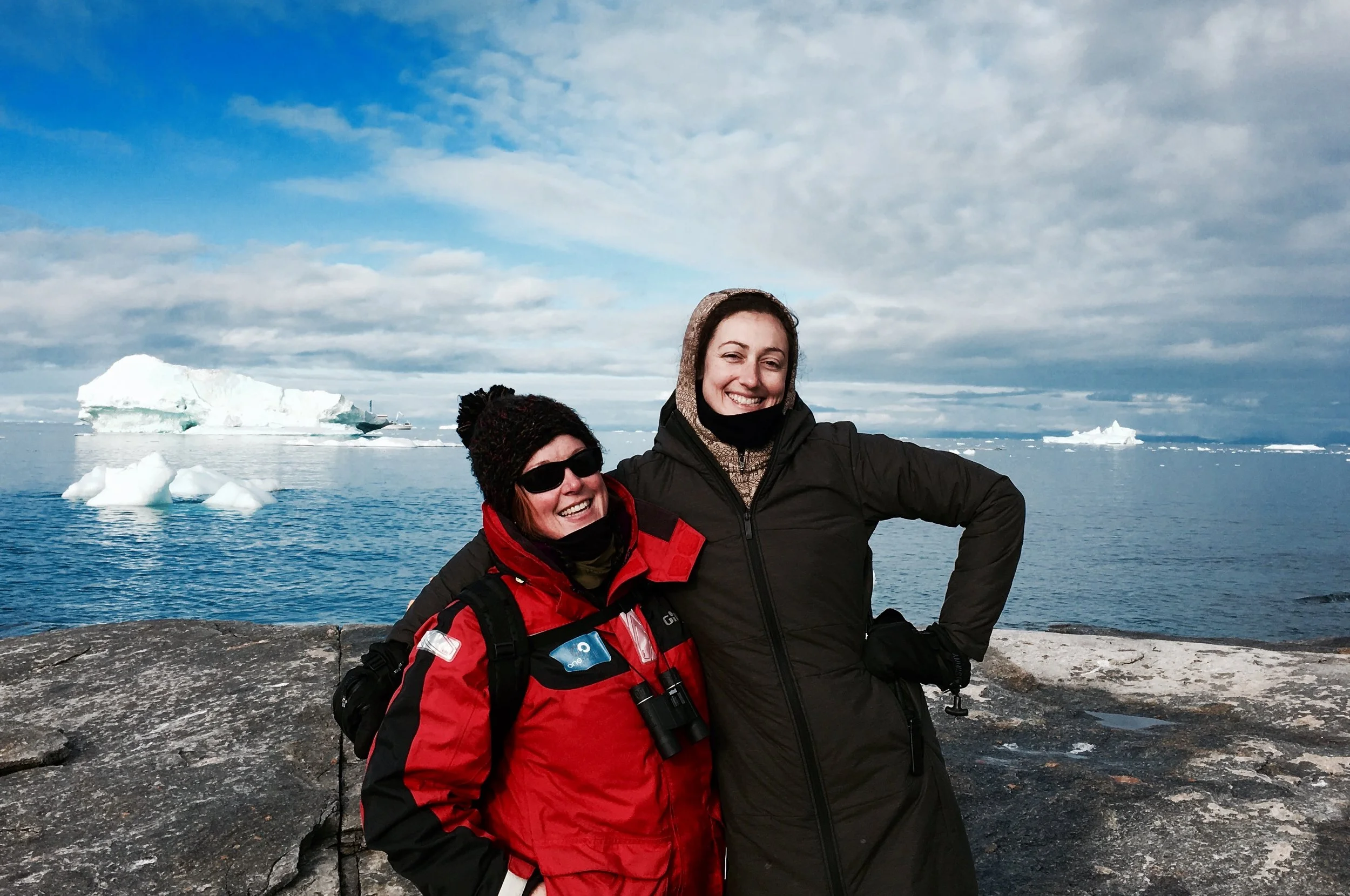Two smiling women in winter jackets on a rocky shoreline with icebergs and ocean in the background.