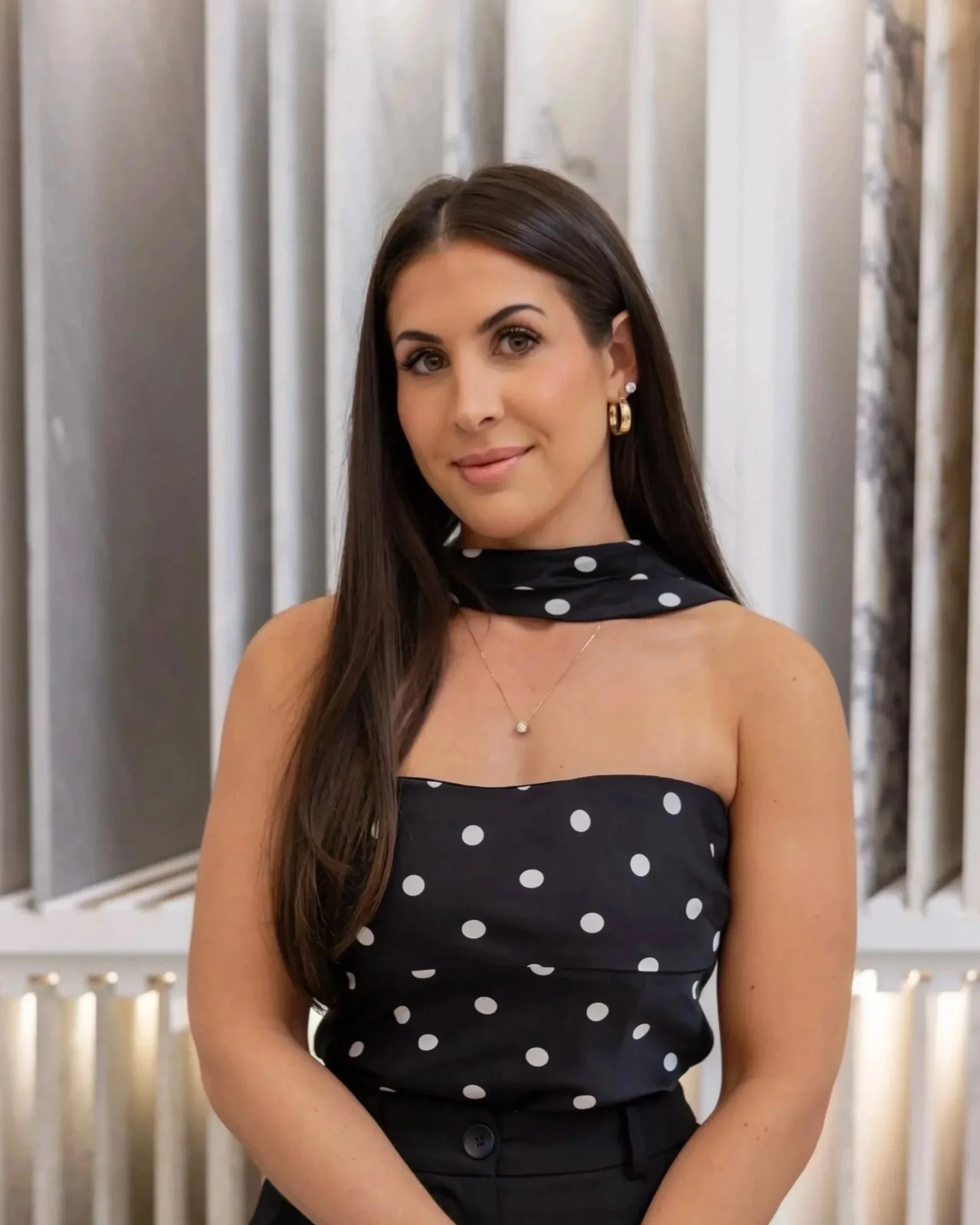 A young woman with long brown hair smiling at the camera, wearing a black top, a gold necklace, and a black jacket, standing in front of a white textured background.