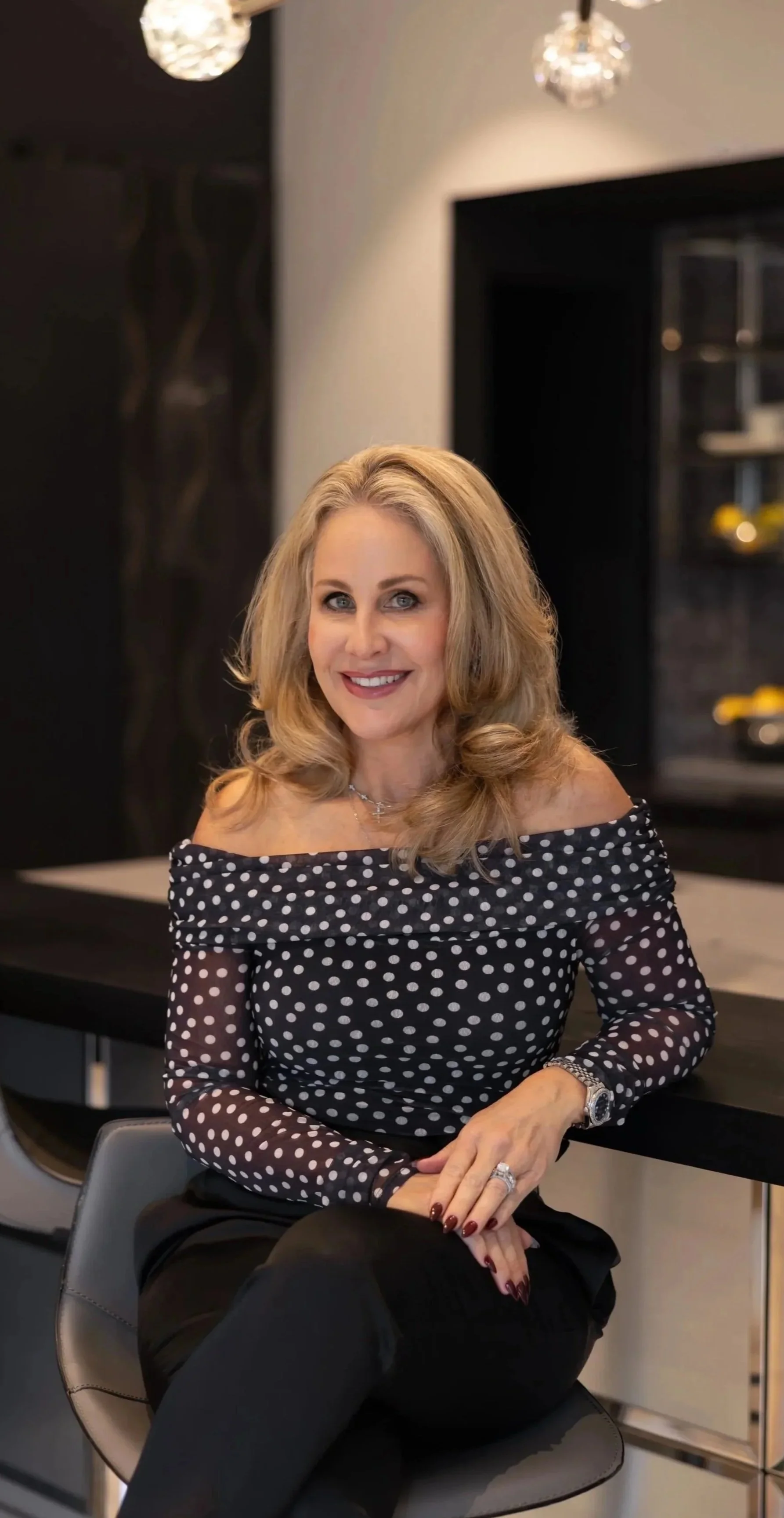 A woman with blonde hair smiling at the camera, indoor setting with a decorative wall in the background.