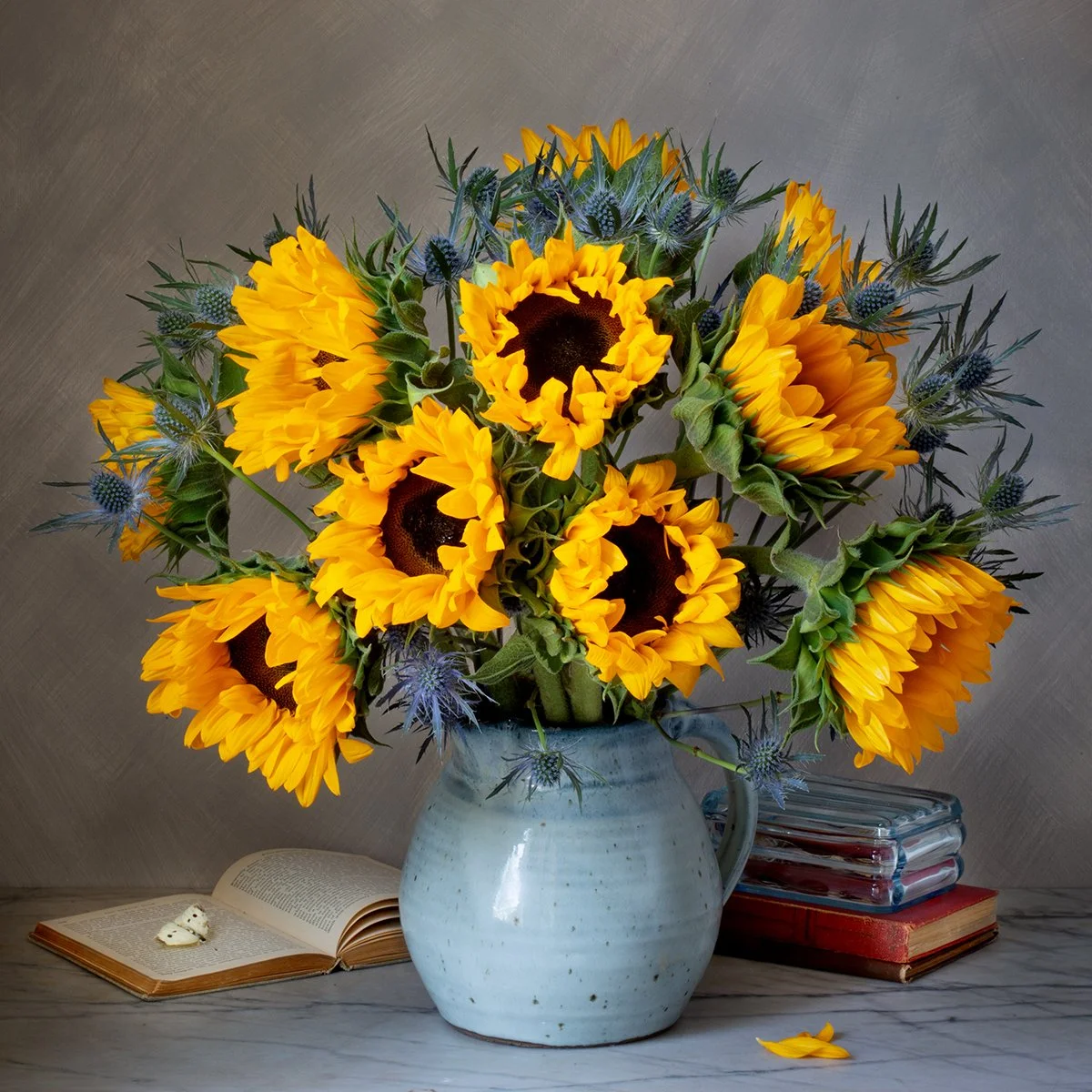 Sunflowers arranged with vintage books and a butterfly in a moody studio still life, lit with soft, dramatic lighting against a dark background.