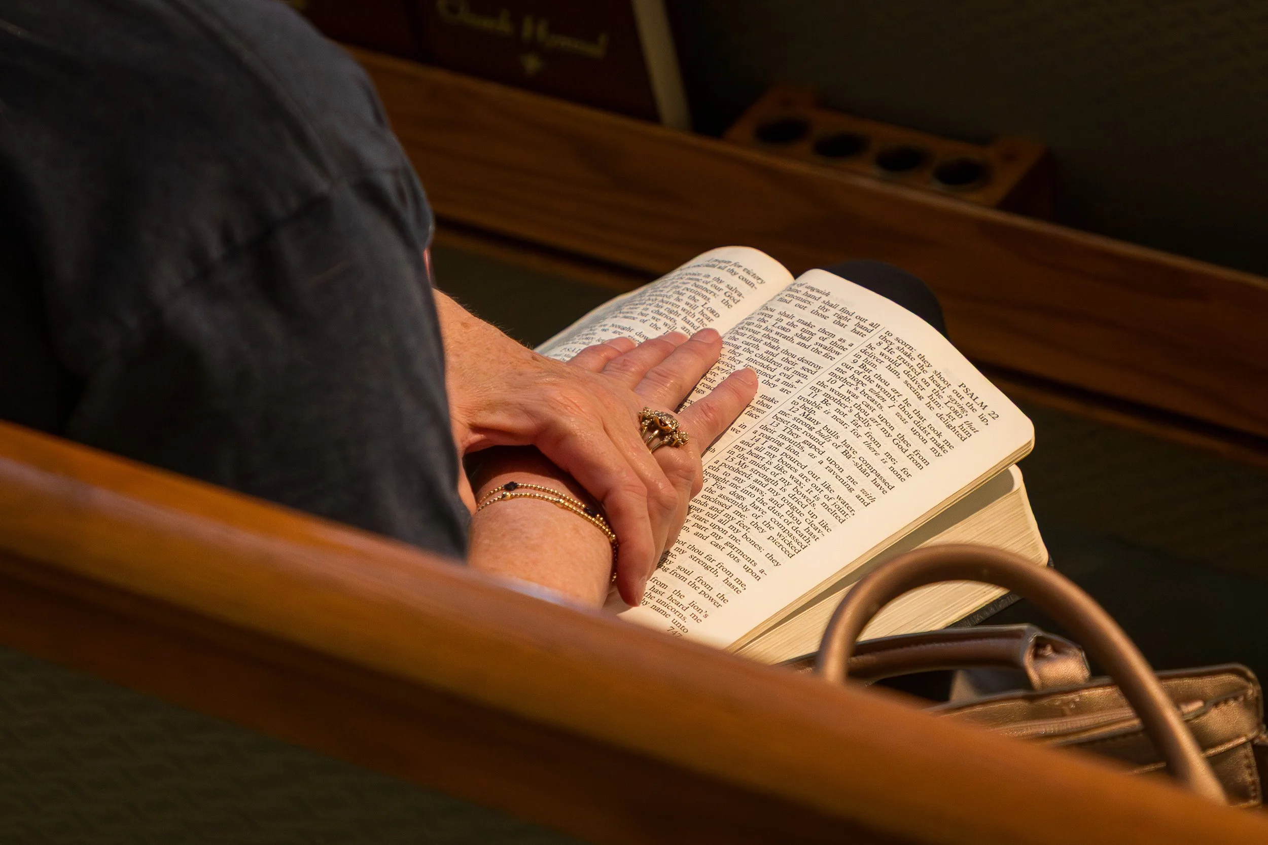 A person reading a Bible in a church pew, with their hand resting on the open pages, wearing rings and bracelets.