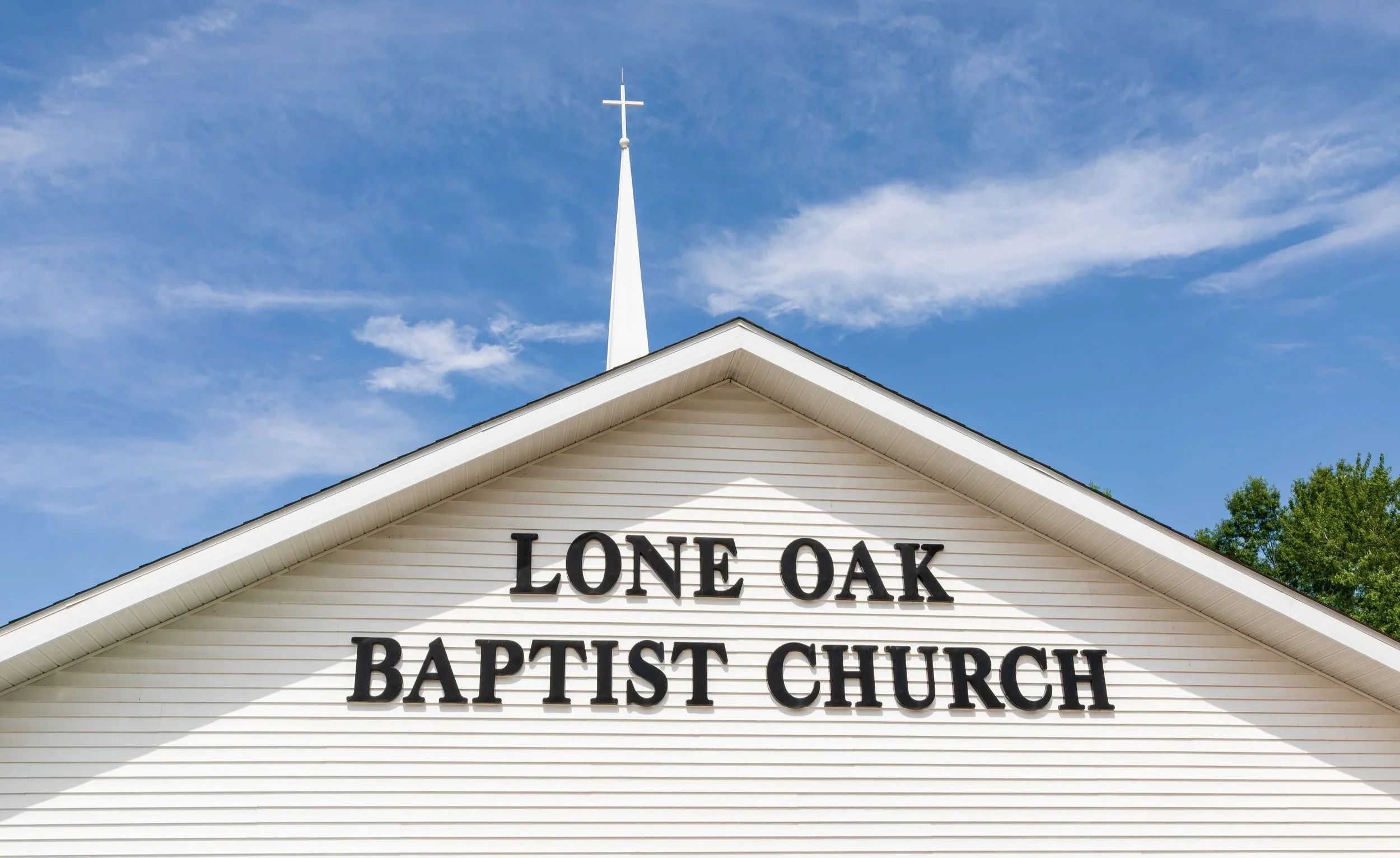 White church with black lettering that reads 'LONE OAK BAPTIST CHURCH' and a steeple with a cross on top, set against a blue sky with some clouds.