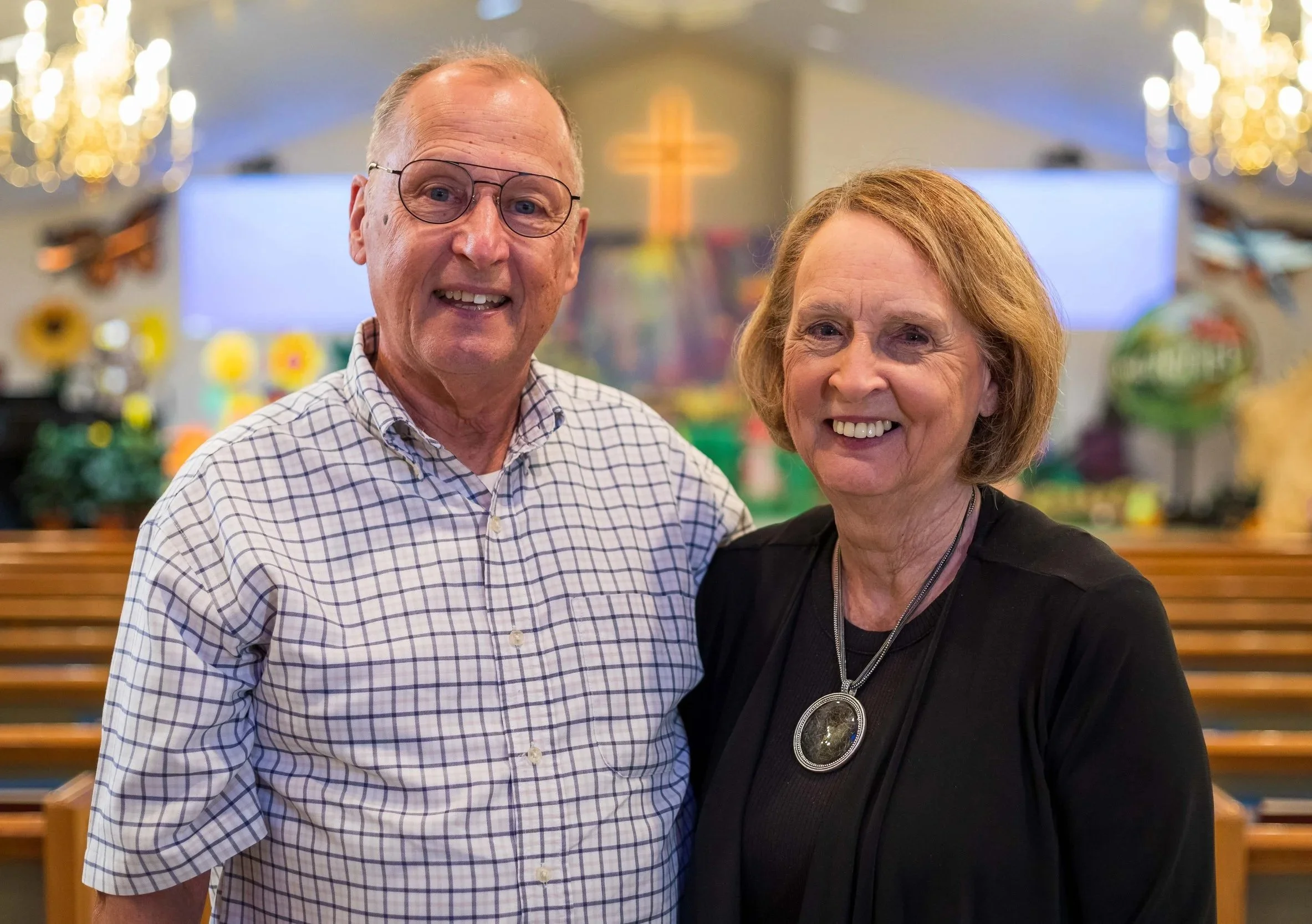 An elderly man and woman smiling inside a church, standing close together with rows of pews behind them and a cross on the wall in the background.