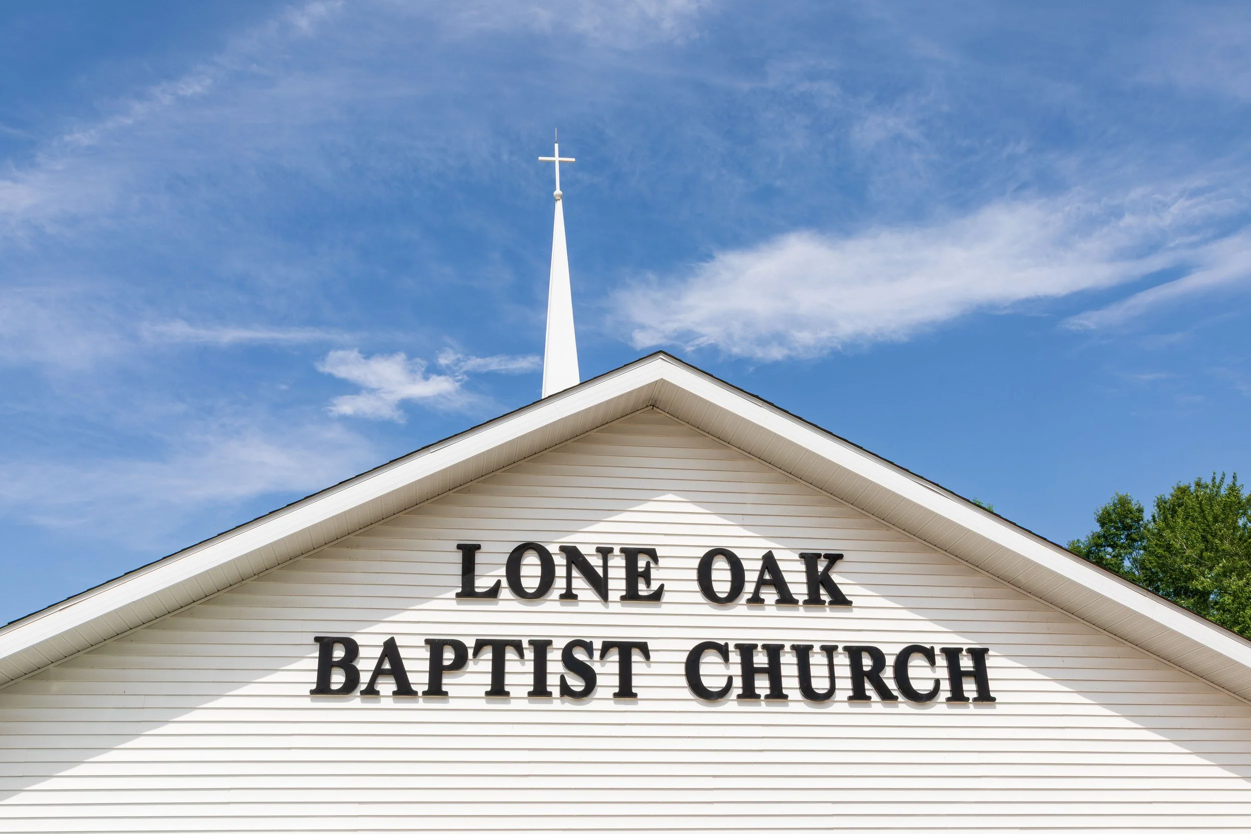 Front of a white church with a steeple and cross, and the sign 'LONE OAK BAPTIST CHURCH' on the facade, under a blue sky with clouds.