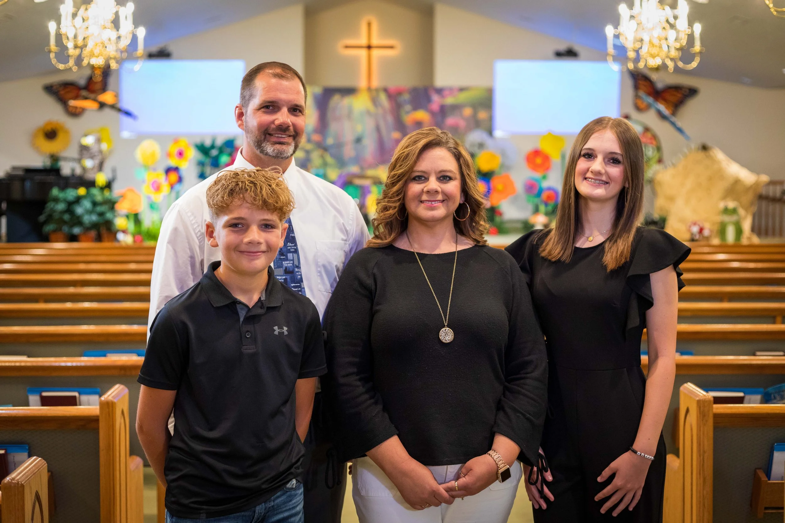 A family of four standing together outdoors by a pond with a fountain, during sunset.